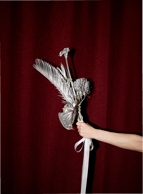 Person holding a bouquet made of metallic or silver-colored decorative elements and feathers against a red curtain background.