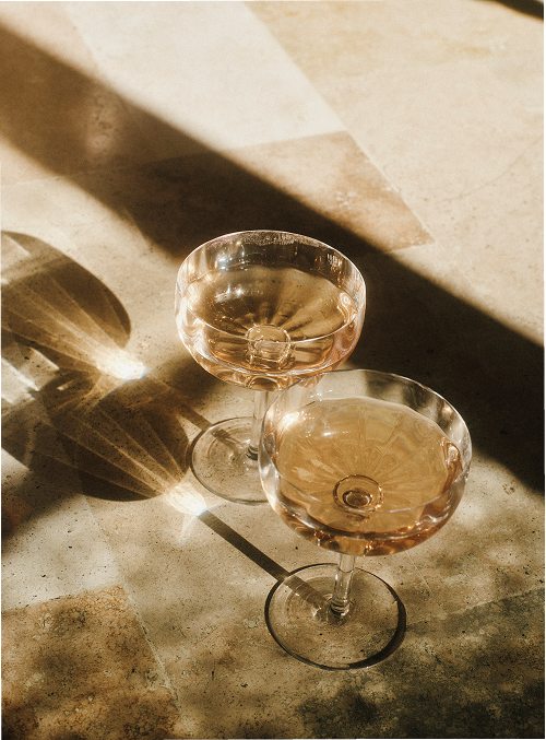 Two coupe glasses filled with rosé wine on a tiled floor with sunlight casting shadows.