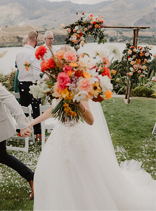 A bride holding a large colorful bouquet of flowers at an outdoor wedding ceremony by a lake with mountains in the background.