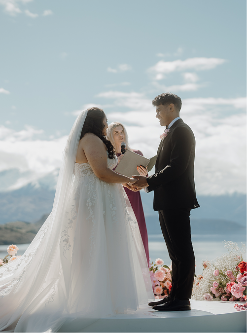 Bride and groom exchanging vows during outdoor wedding ceremony with officiant, mountain and lake backdrop, and floral decorations.