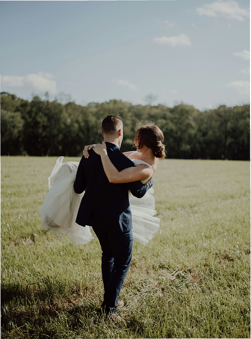 A couple in formal wedding attire, with the groom carrying the bride in his arms in an open grassy field with trees in the background and a partly cloudy sky.