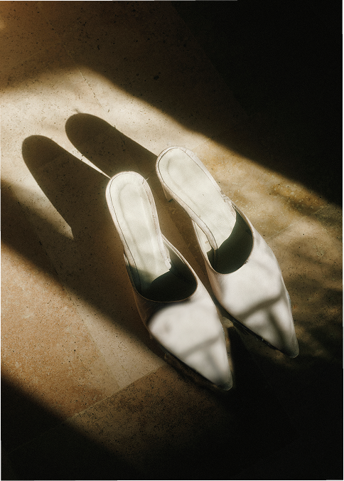 Pair of white high-heeled shoes on a beige surface, with shadows cast from sunlight.