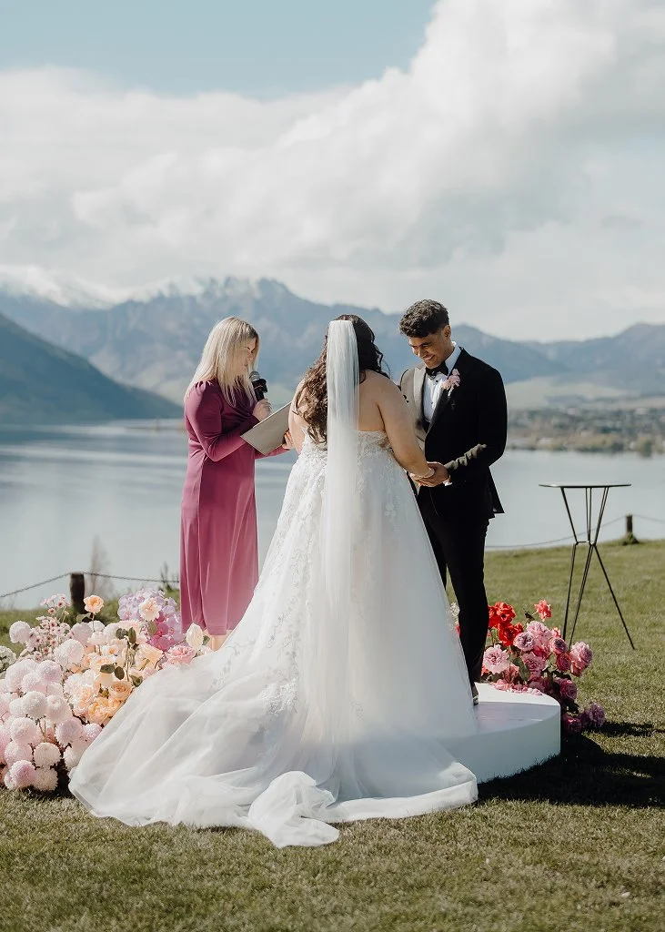 A couple getting married outdoors by a lake with mountains in the background, exchanging vows while a woman officiant stands nearby.