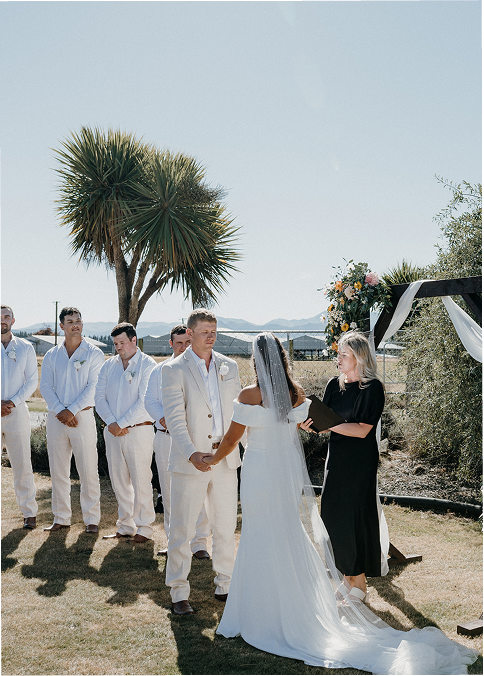 Bride and groom exchanging vows during an outdoor wedding ceremony with groomsmen and officiant present, under a clear sky with desert and palm trees in the background.