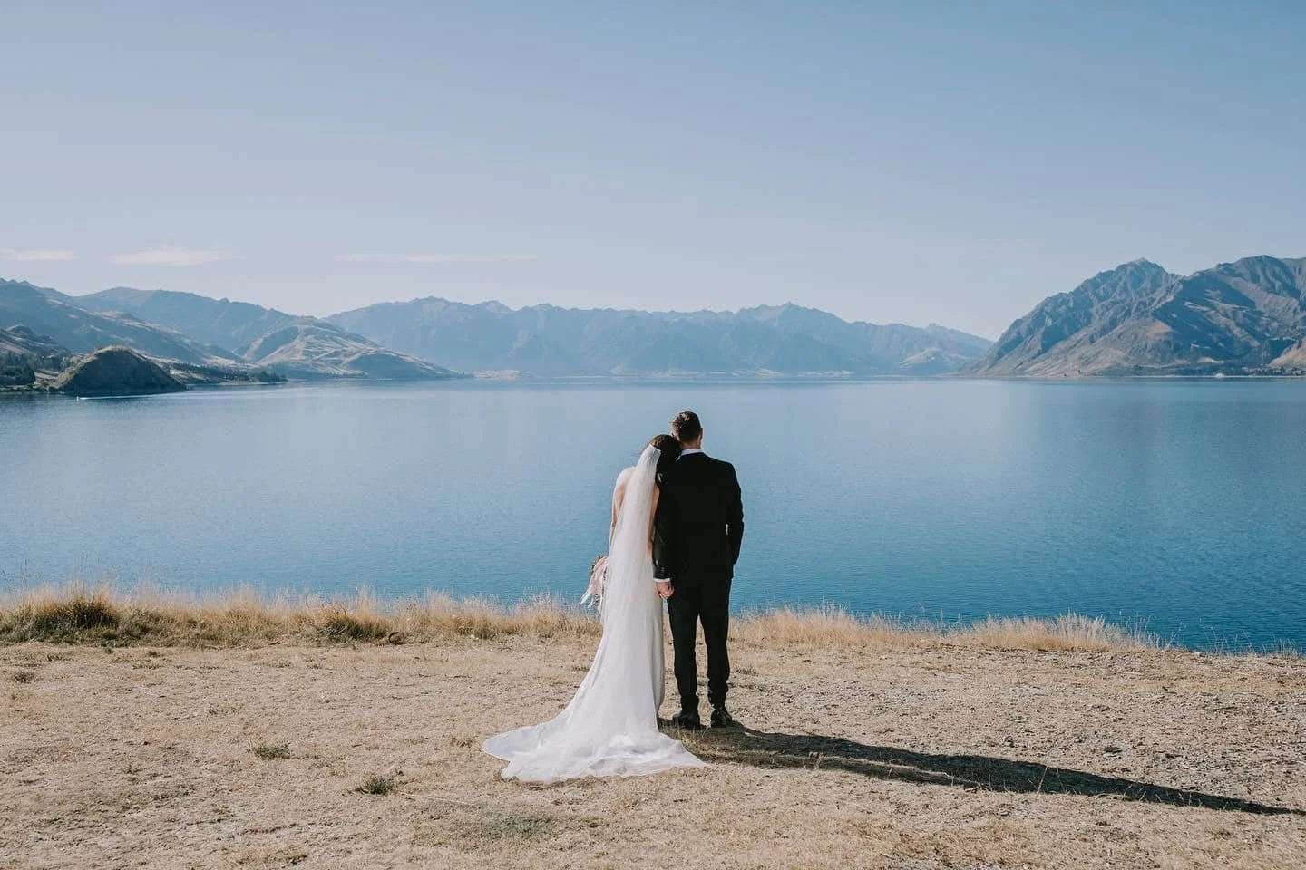 A bride and groom stand on a dirt path by a large lake, mountains in the background, with clear blue skies overhead