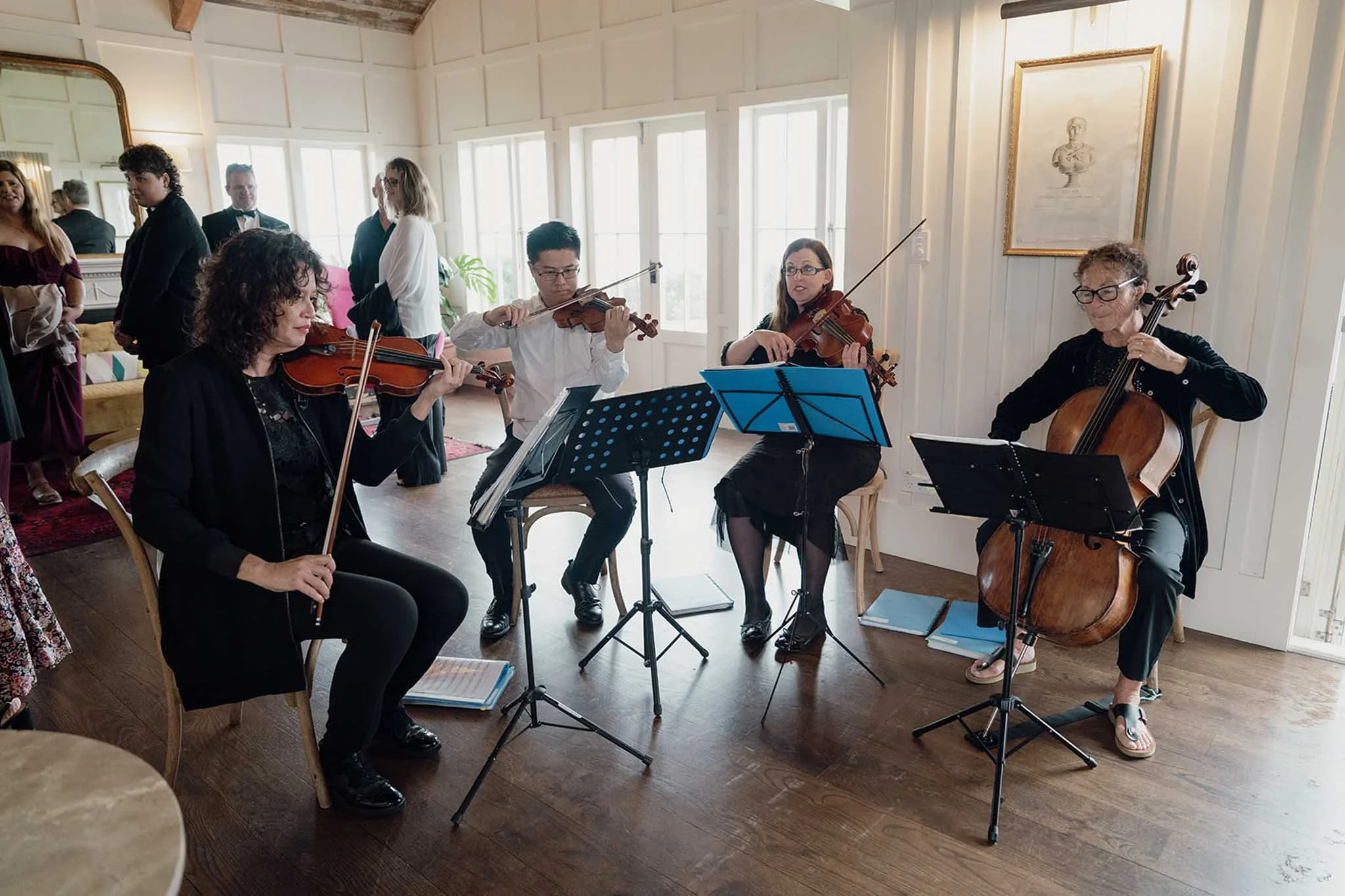 A group of musicians playing string instruments, including violins and a cello, in a bright, airy room with large windows and wooden flooring. Some guests are standing in the background, observing.