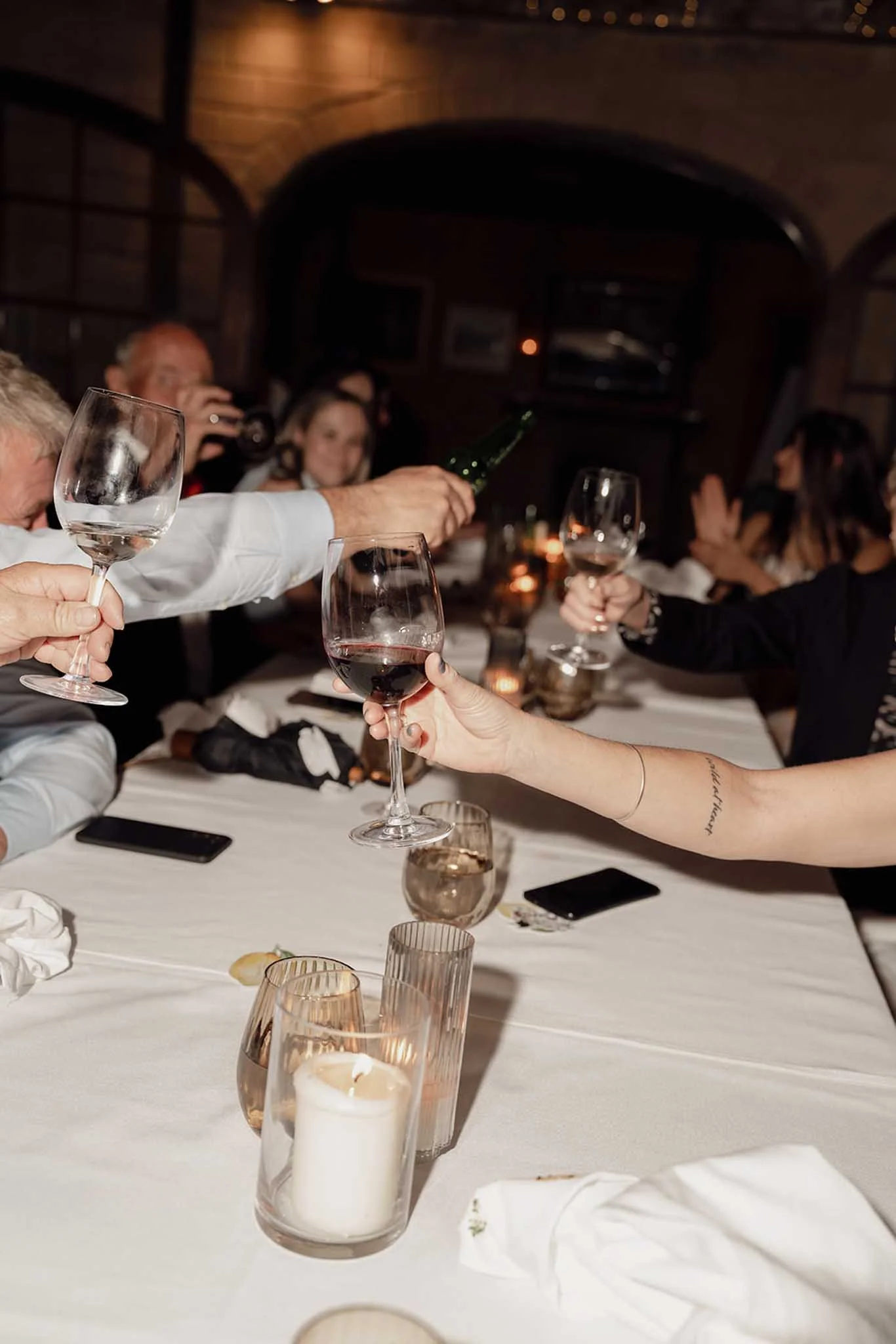 People raise glasses of red wine in a toast around a long, elegantly set dinner table in a dimly lit restaurant or banquet hall. Candles and a white tablecloth are visible.