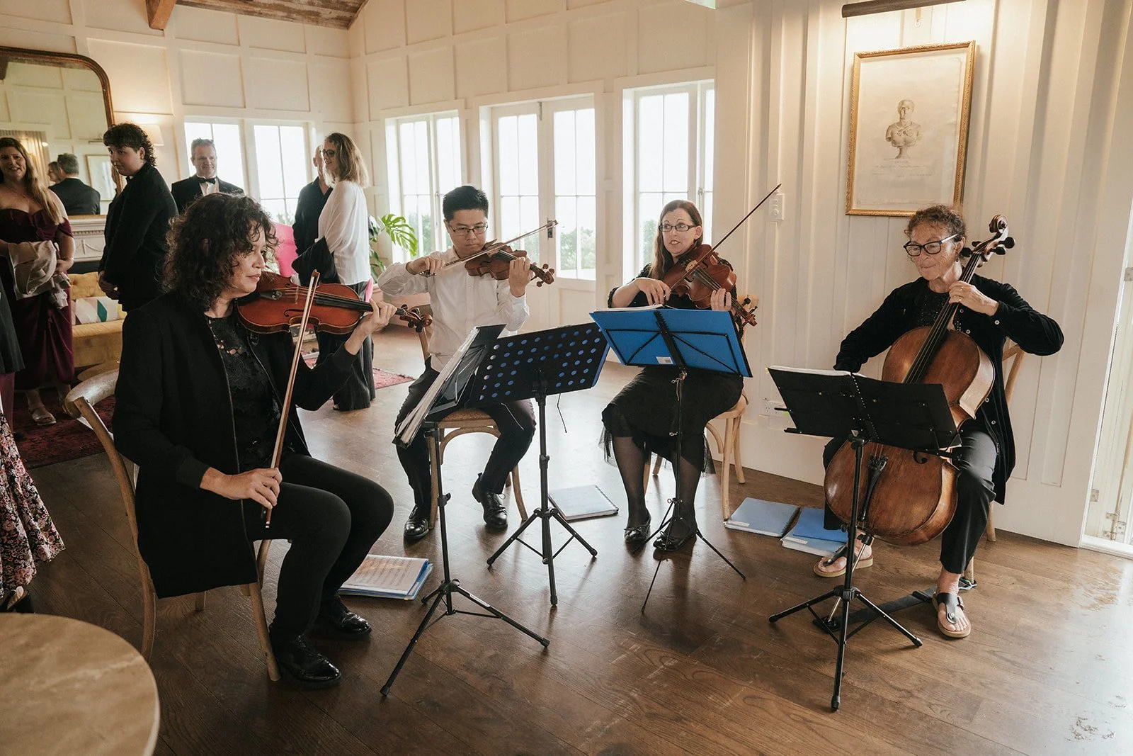 A group of five musicians playing string instruments, including violins, viola, and cello, in a cozy, well-lit room with large windows and wooden floors during a performance or rehearsal.