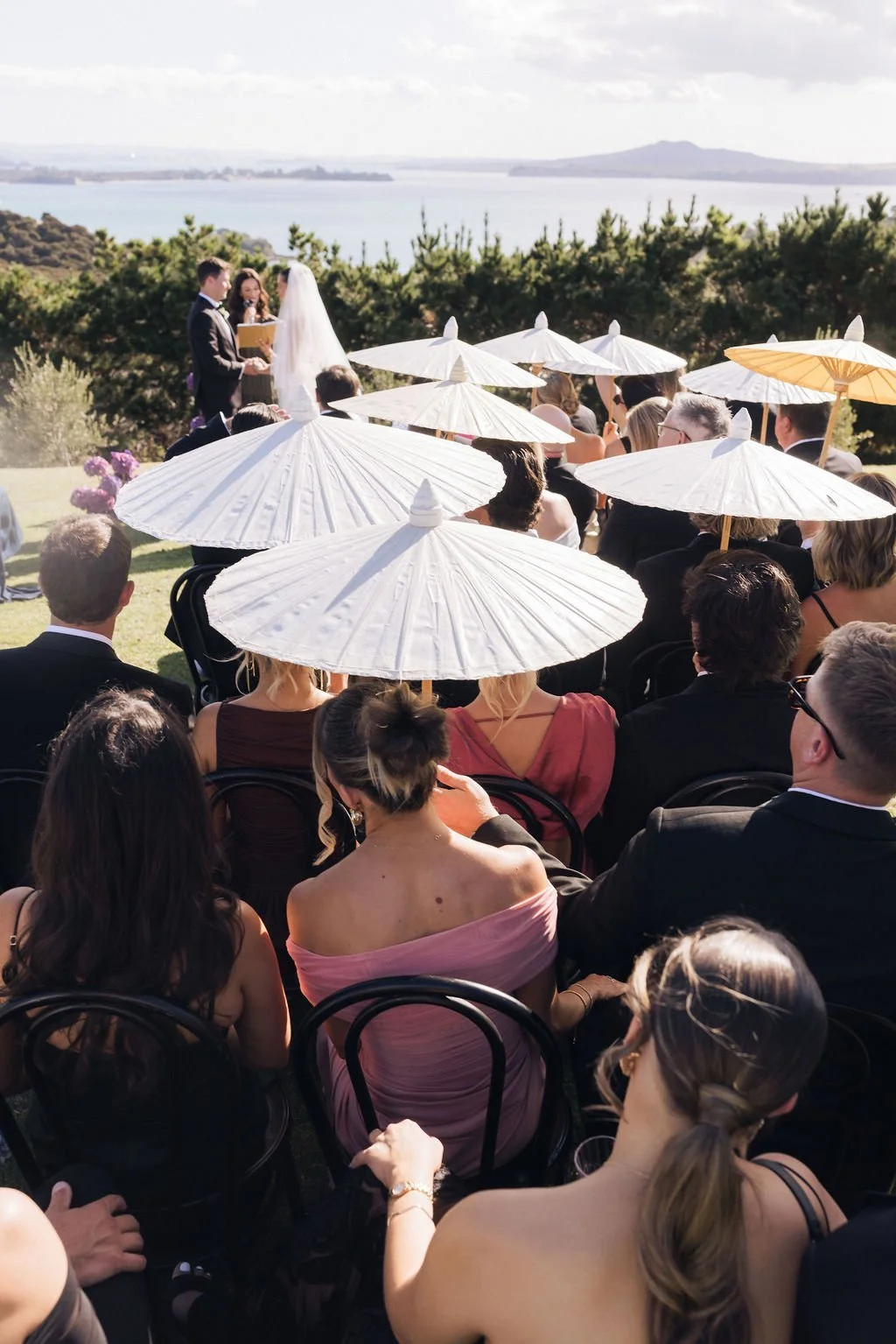 An outdoor wedding ceremony with guests seated under white umbrellas, watching a couple exchange vows by a lake with hills in the background.