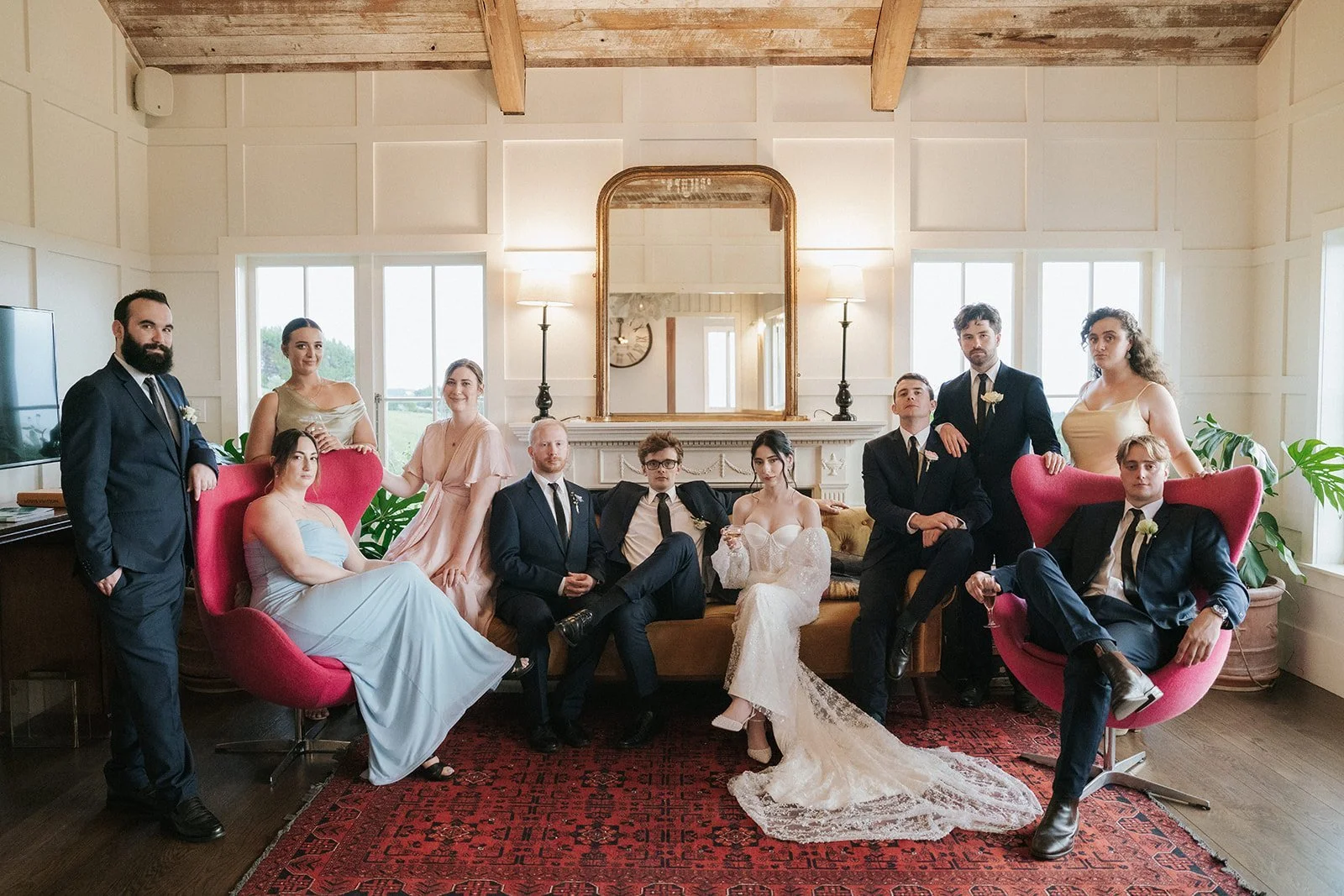 Group of people dressed in formal attire posing in a stylish living room with vintage furniture, large windows, and a decorative mirror.