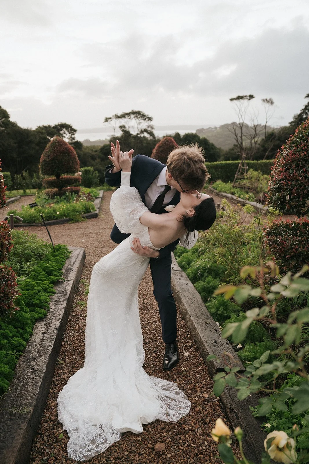 A newlywed couple is in a garden, with the groom holding the bride in a dip and sharing a kiss, surrounded by lush greenery and topiary bushes.