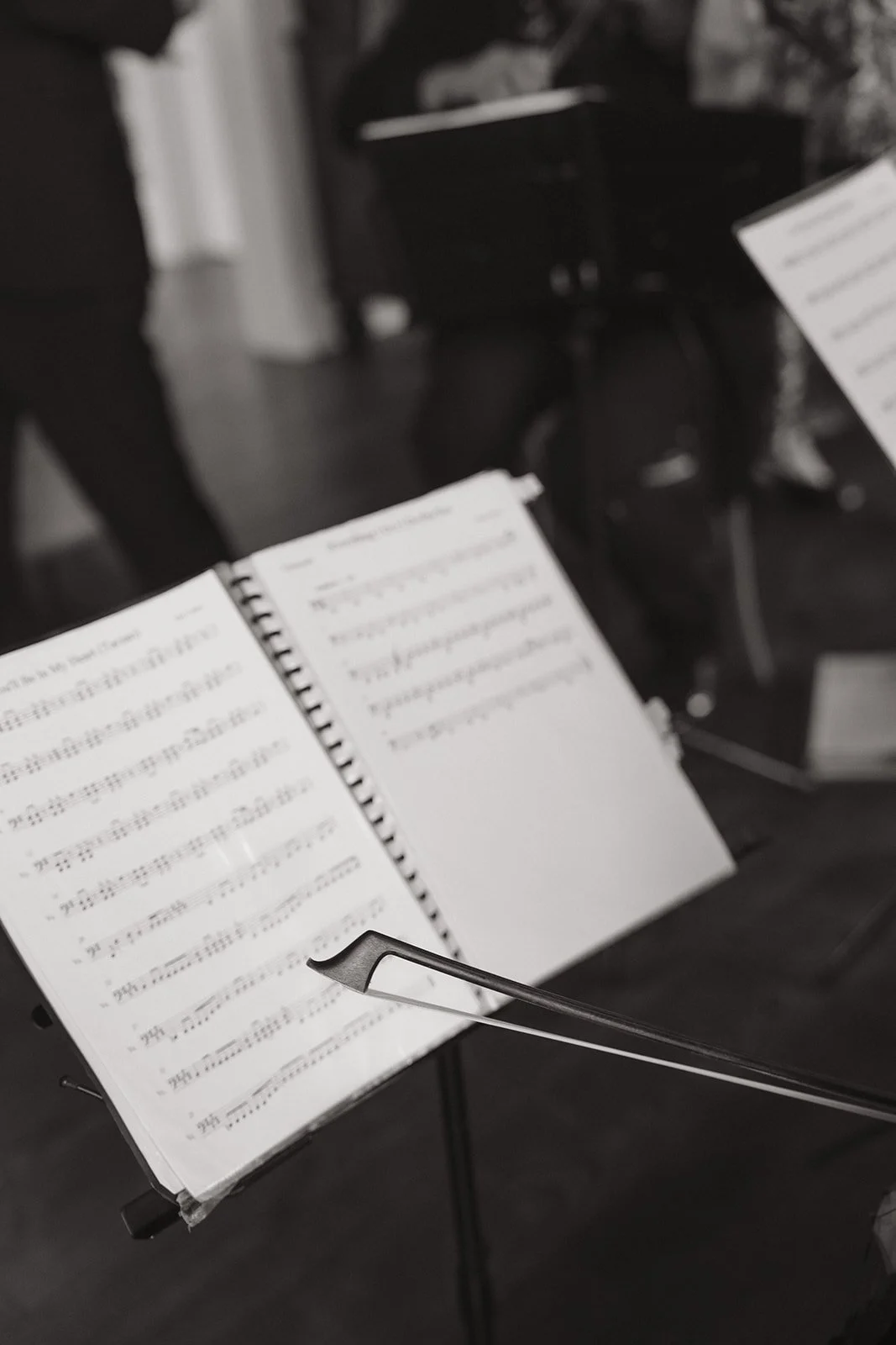 An open sheet music stand with musical notes and sheet music, a baton lying on the stand, and people and chairs in the background in a dark room.
