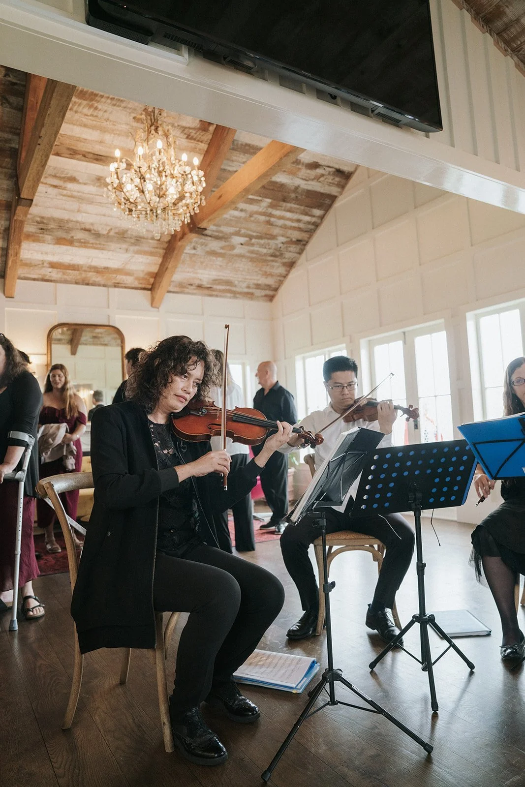 Musicians playing violins during a performance in a bright, rustic room with a chandelier and large windows.