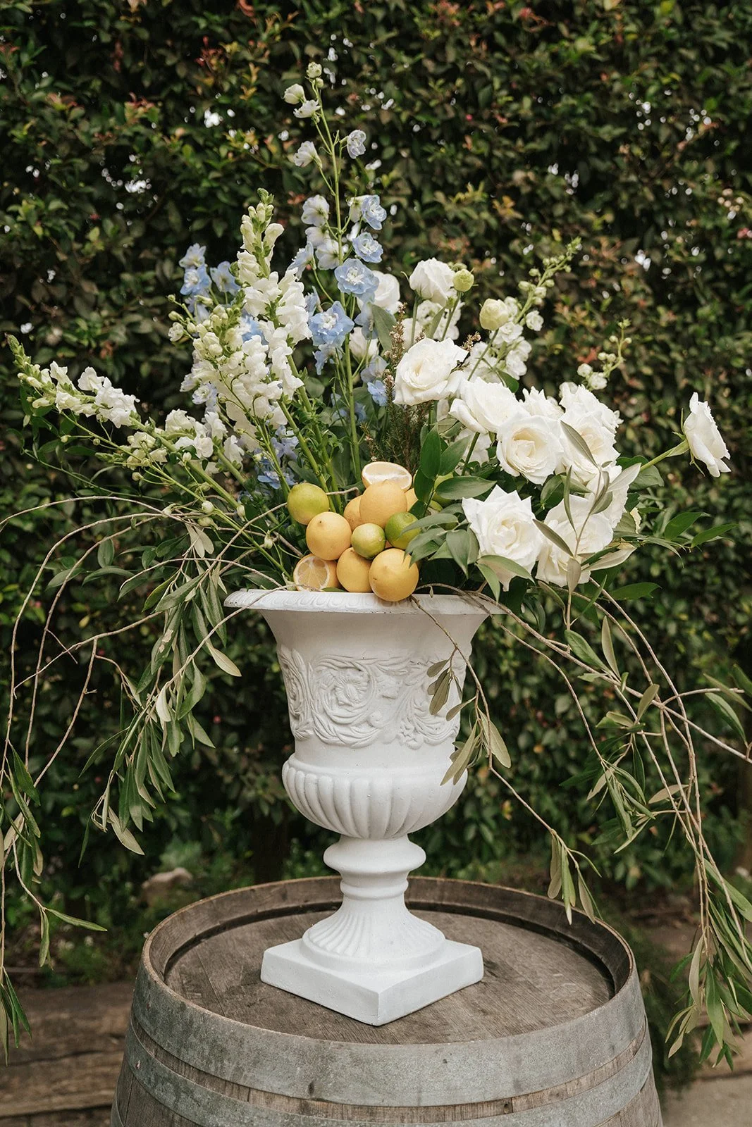 A decorative white urn filled with white and light blue flowers, lemons, and greenery, placed on a wooden barrel outdoors.