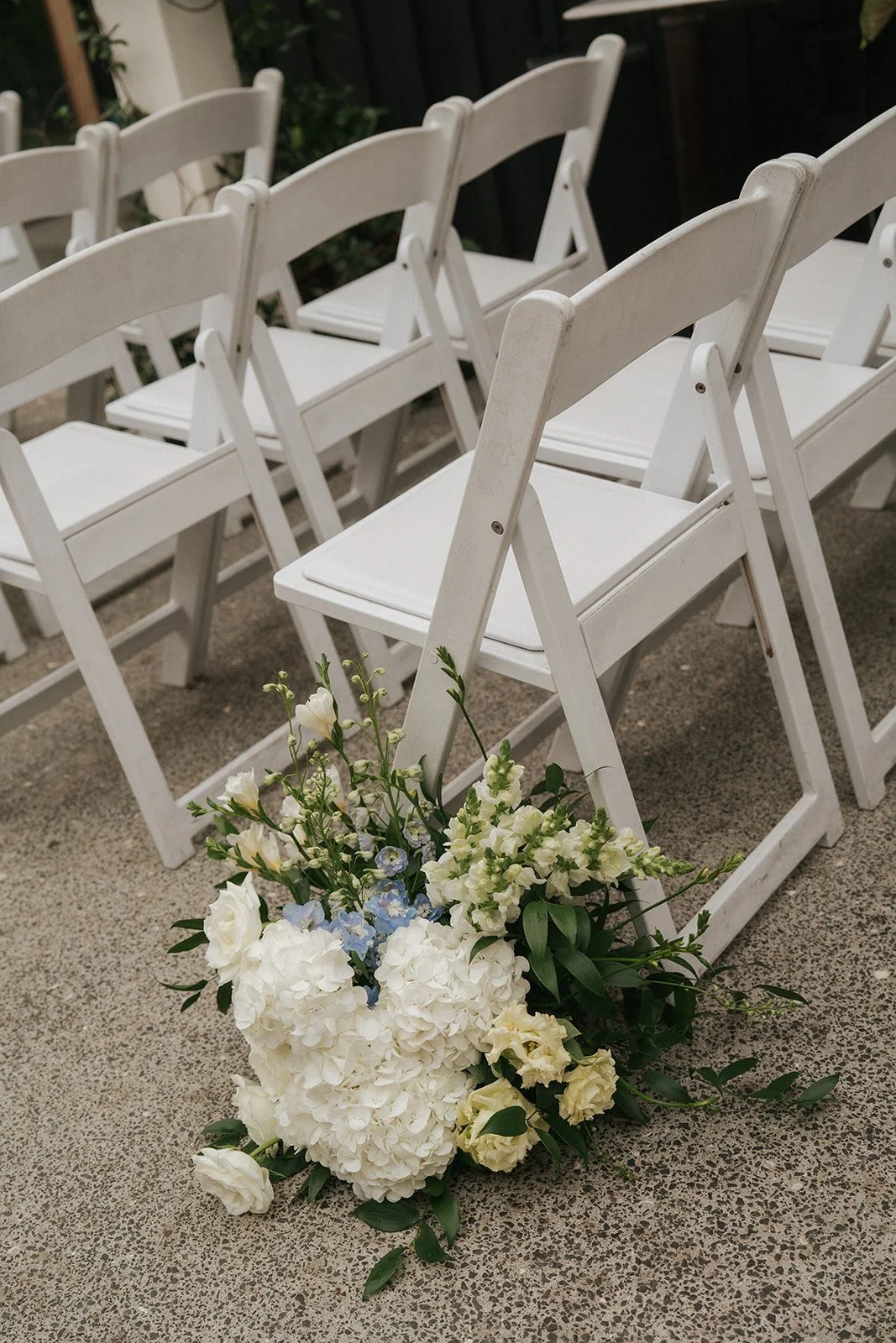 A row of white folding chairs arranged on a concrete floor, with a flower arrangement of white hydrangeas, lisianthus, and blue and green accents placed at the end of the row.