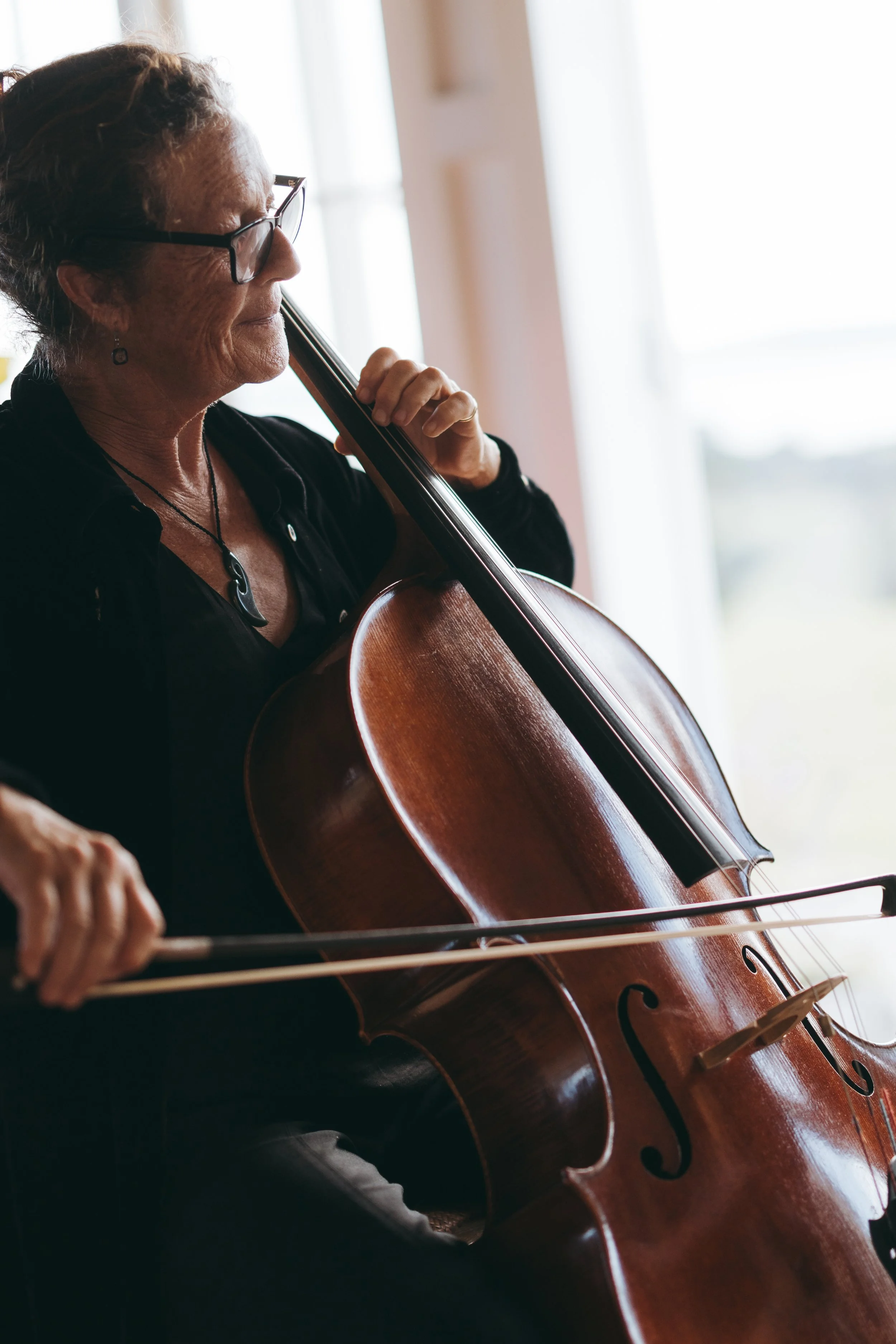 Elderly woman with glasses playing a cello, focusing on her face and instrument in a bright room.