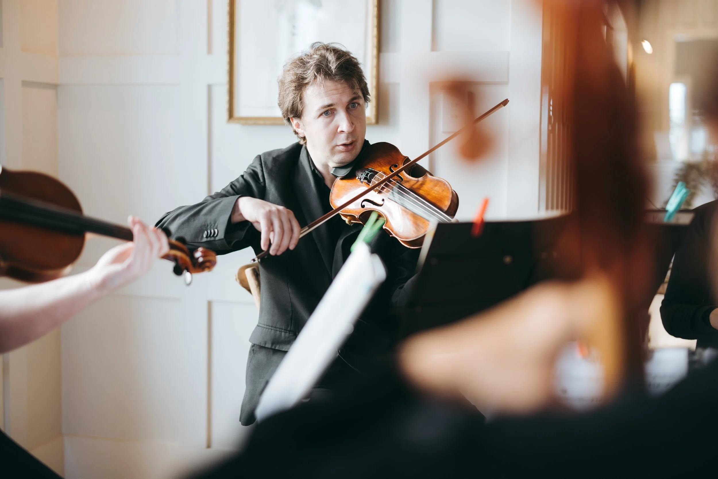 A male violinist performing during an indoor concert, with a focused expression, surrounded by other musicians and sheet music.