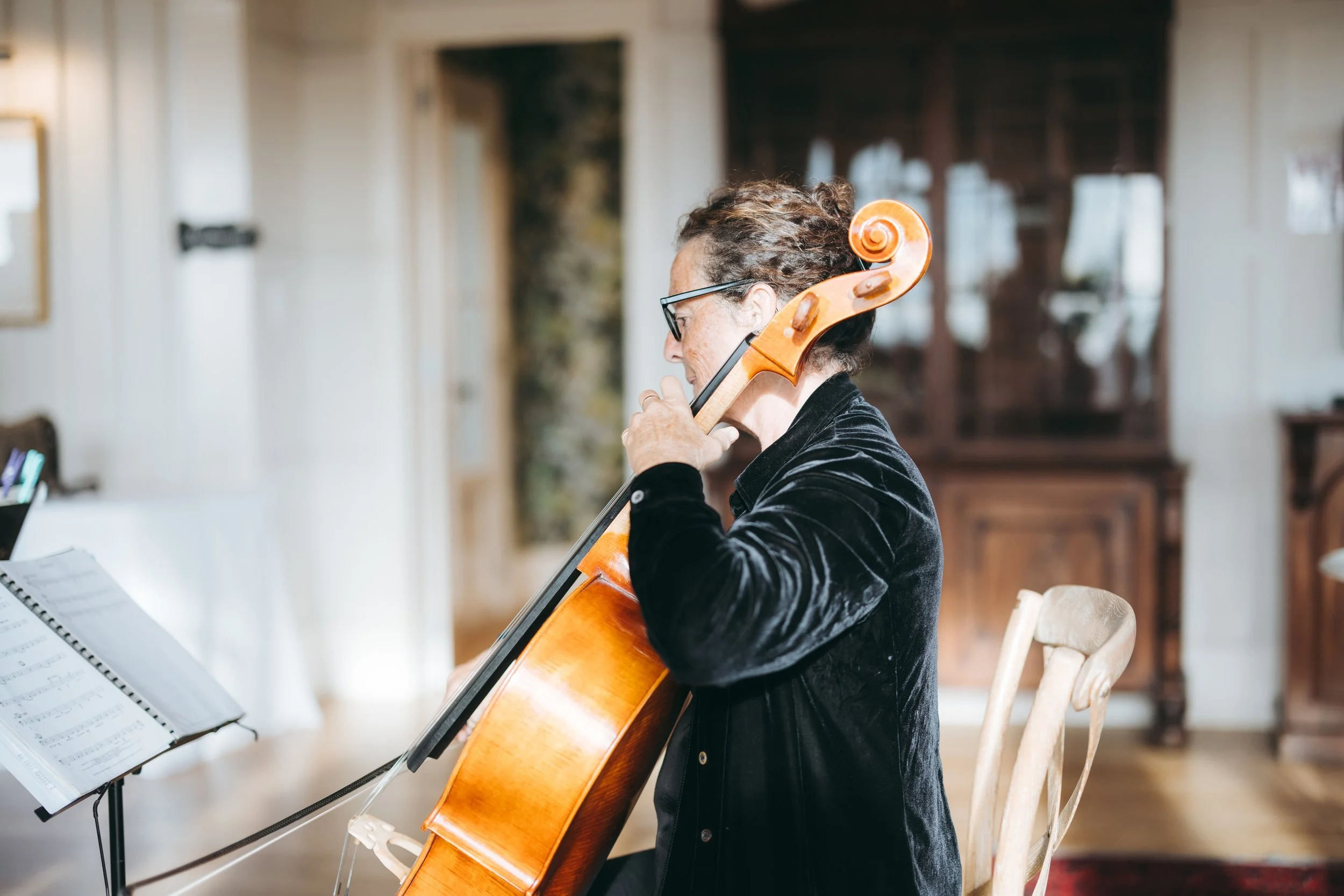 A woman playing a cello indoors, sitting on a wooden chair with sheet music on a stand nearby.