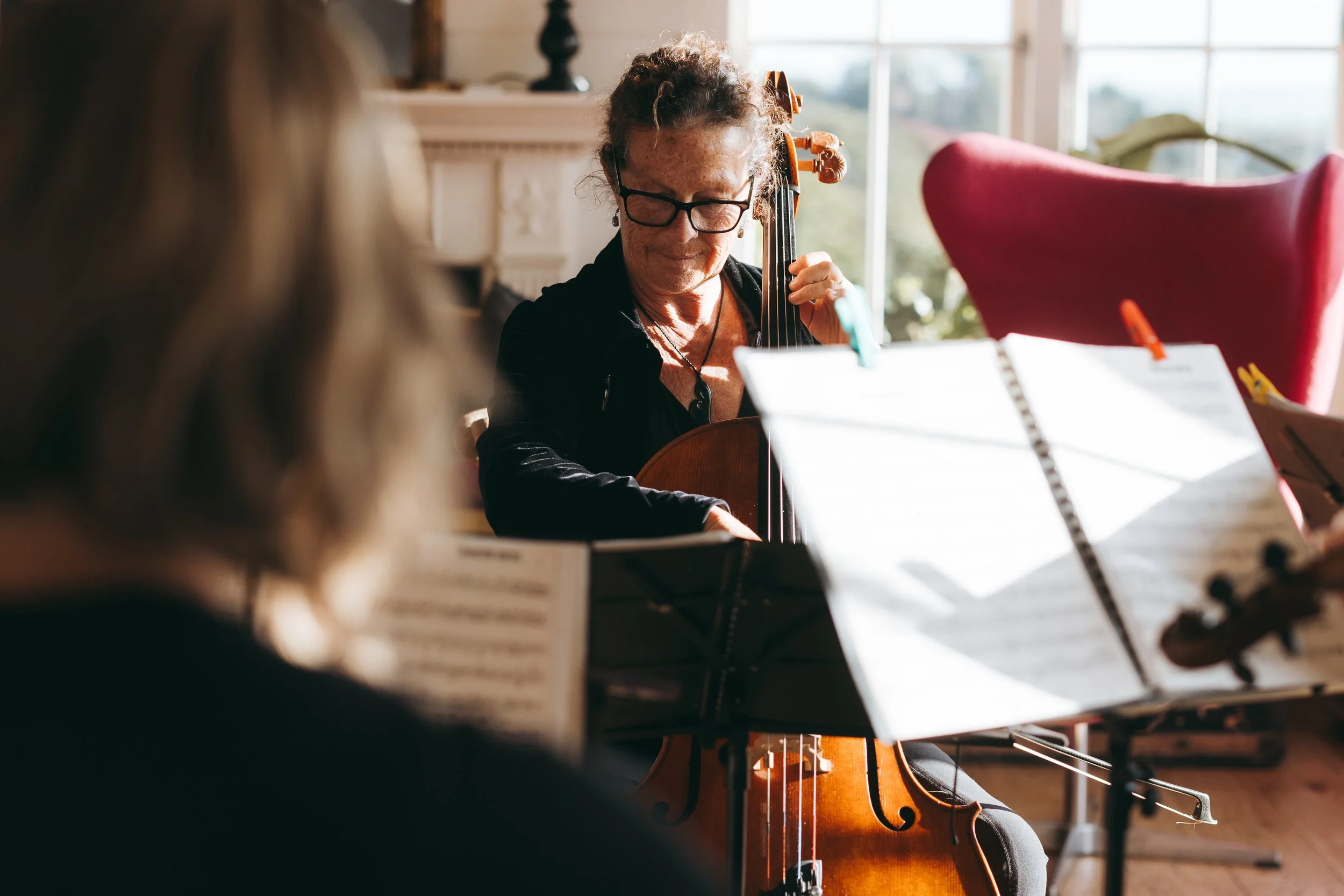 A woman with curly hair, glasses, and earrings playing a cello during a music practice or lesson, with sheet music and another person in the foreground.