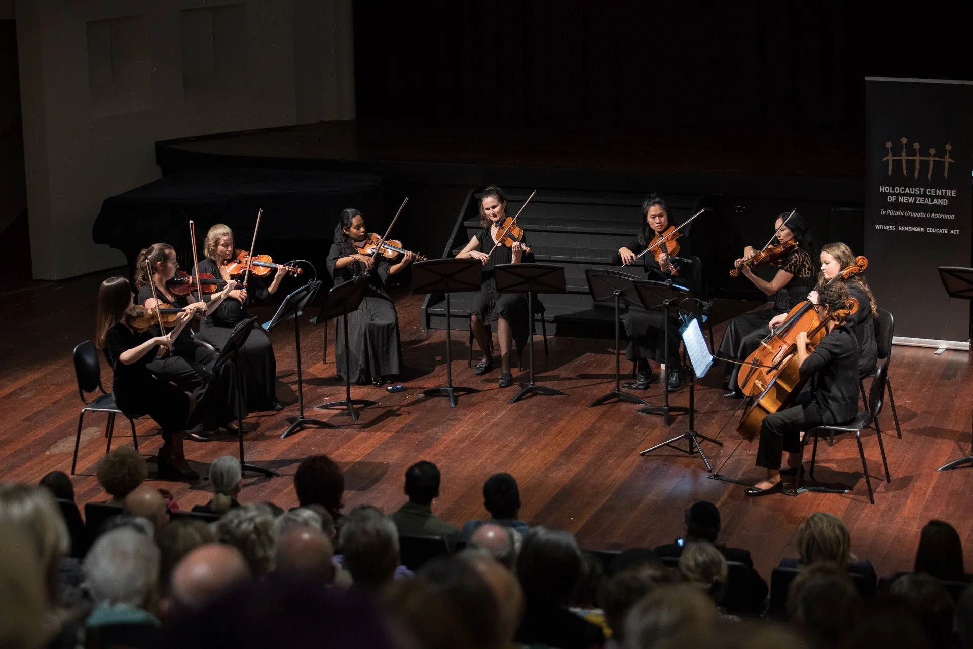 Orchestra of nine women playing violins and cellos on stage during a concert in front of an audience