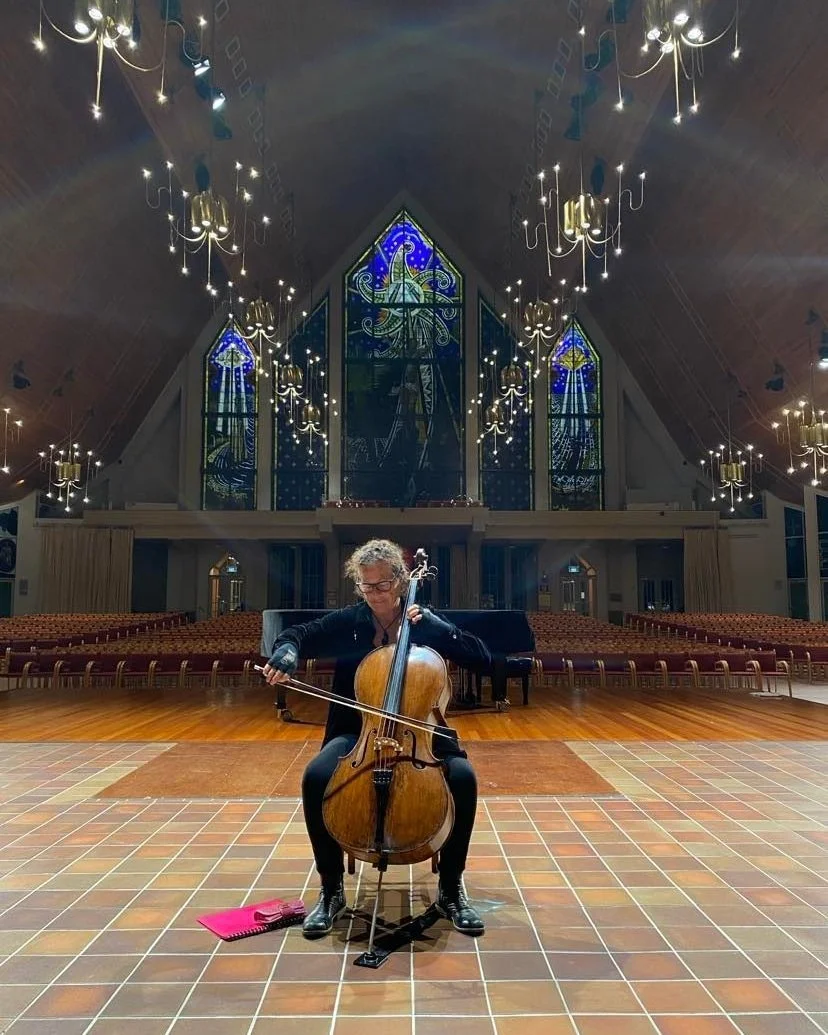 A woman playing the cello on a stage inside a church with stained glass windows and chandeliers overhead.