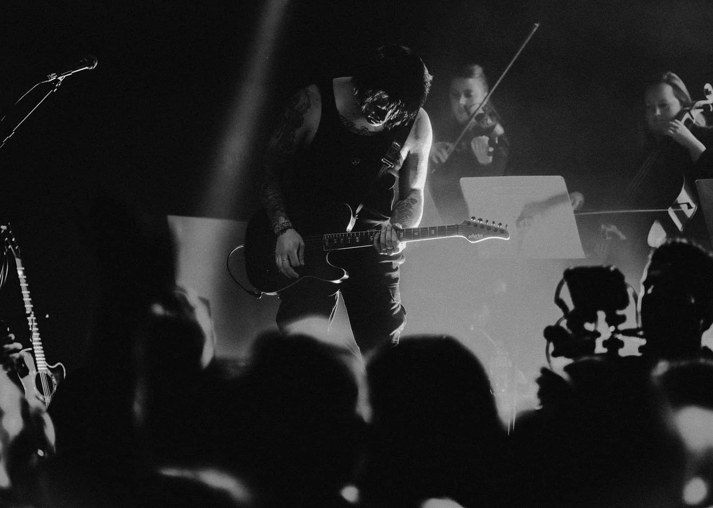 Black and white photo of a musician playing an electric guitar on stage, with two violinists in the background, and a crowd in front of the stage.