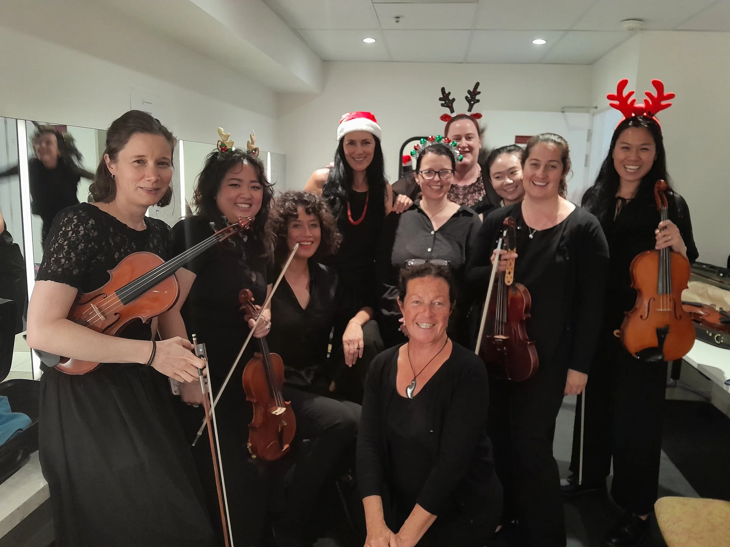 Group of women gathered together indoors, dressed in black, some wearing Christmas headbands and accessories like reindeer antlers and wreaths, holding musical instruments such as violins. It appears to be a festive holiday gathering.