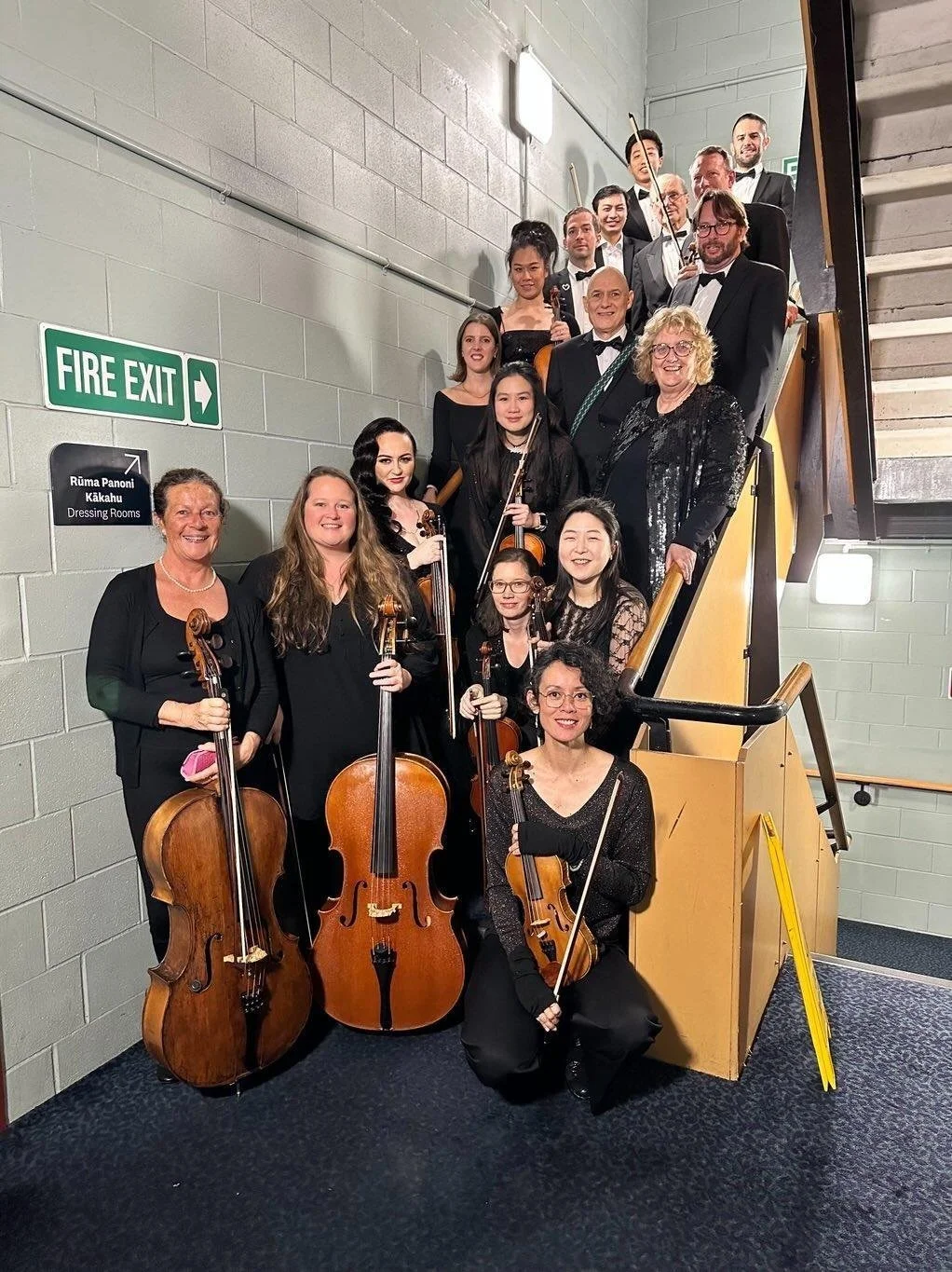 Orchestra group photo on a staircase, with string instruments, dressed in formal black attire, inside a building with gray walls and a fire exit sign.