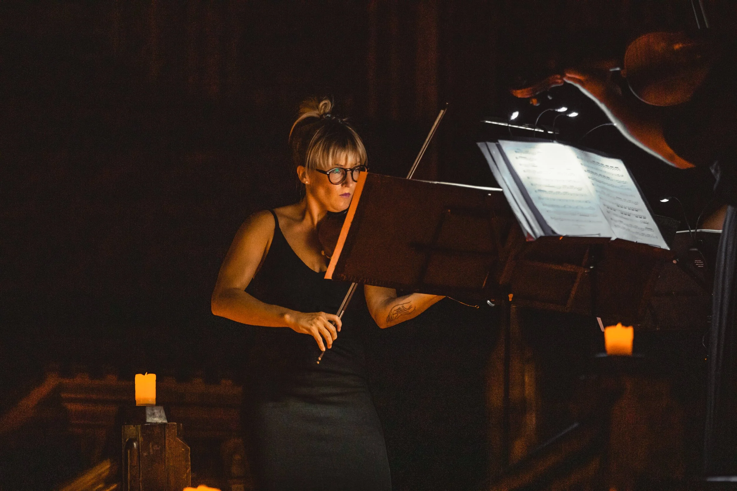 A woman with glasses playing the piano in a dark, intimate setting illuminated by candles.