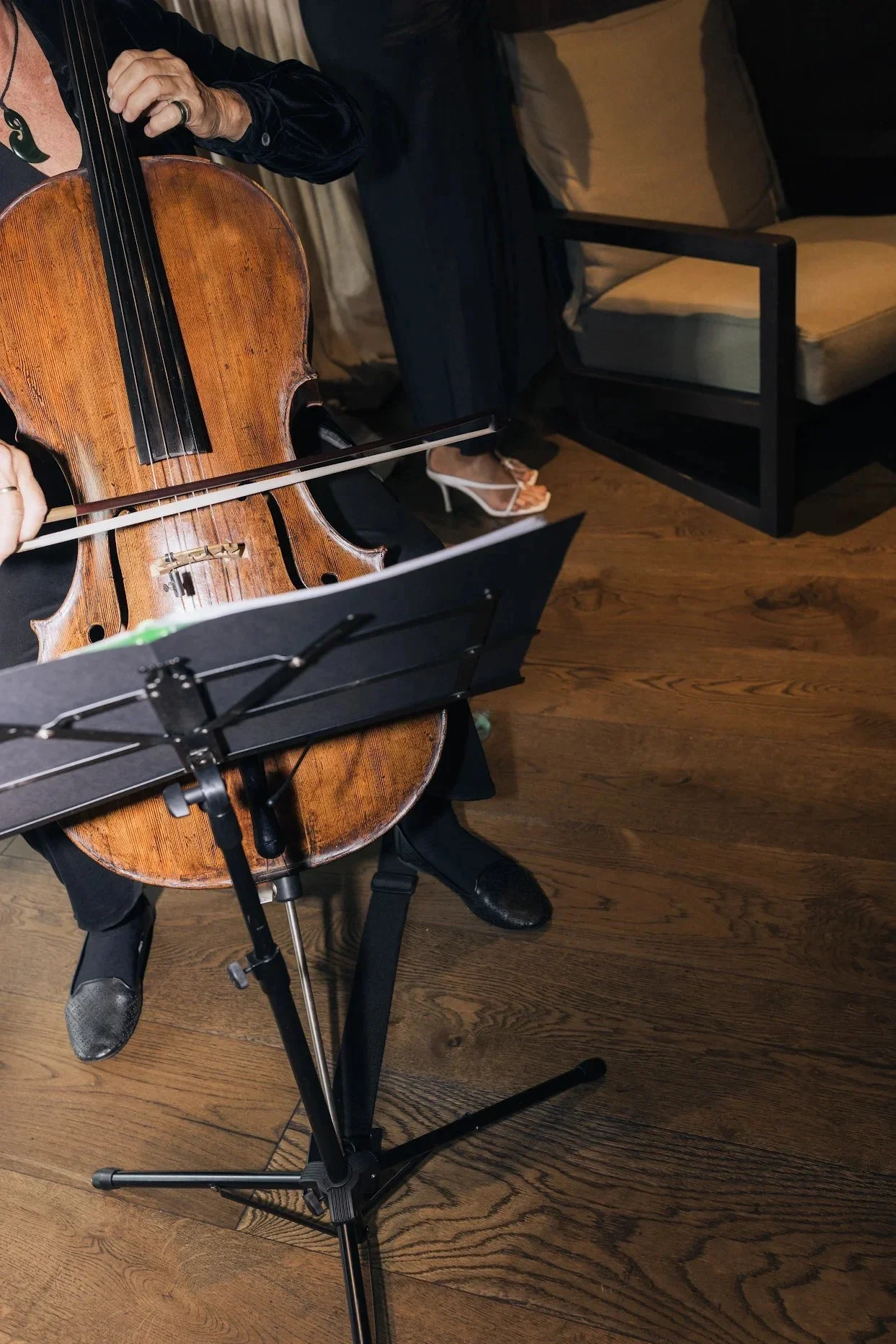 A person playing a cello, with a music stand in front. The person is wearing black pants, black shoes, and a partially visible black jacket. The scene is indoors with wooden flooring, a chair, and a beige couch in the background.