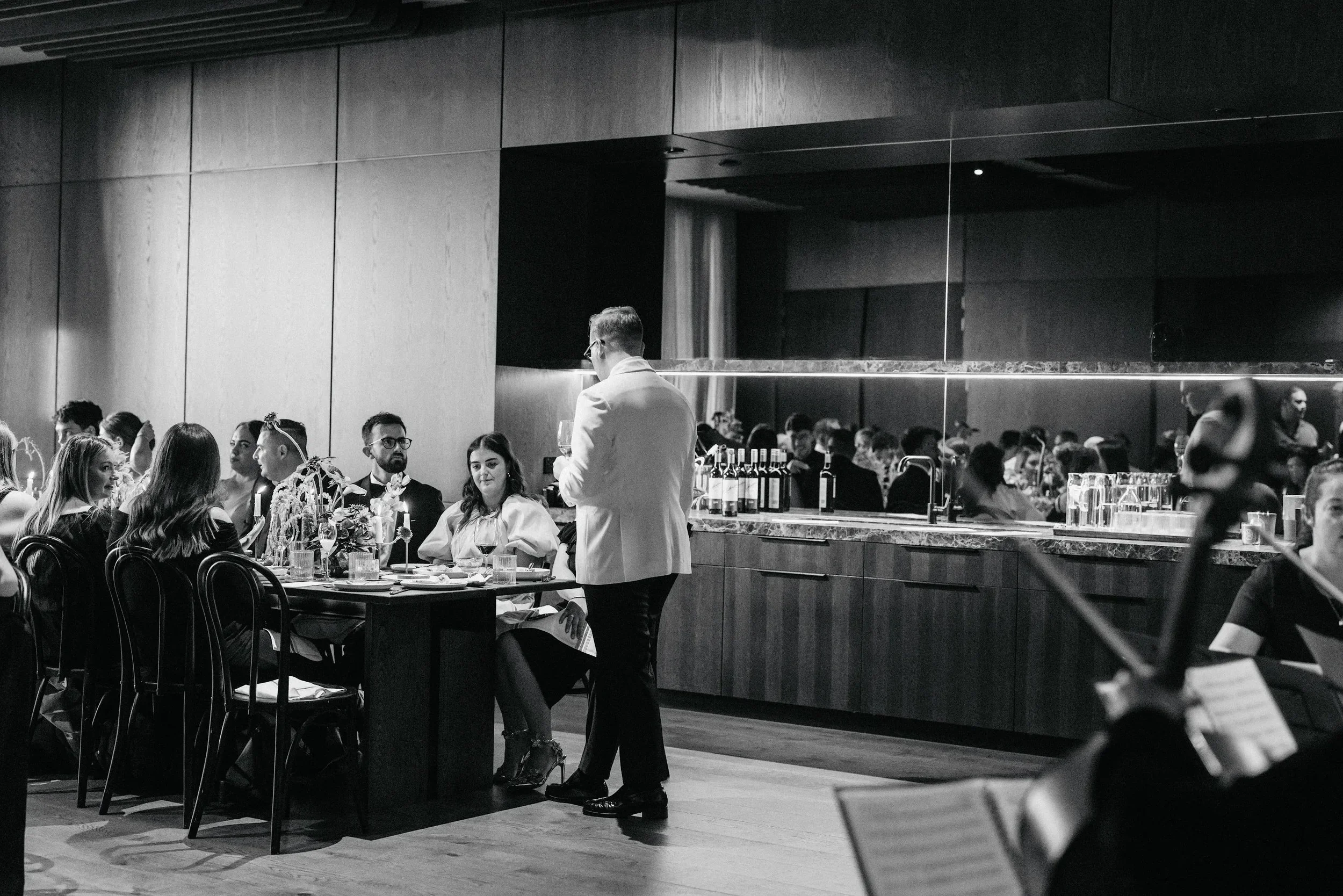 A black and white photo of a restaurant or event space with people seated at tables and a server in a white jacket taking an order. The background shows a bar with bottles and glasses, and the space has modern wooden walls and ceilings.