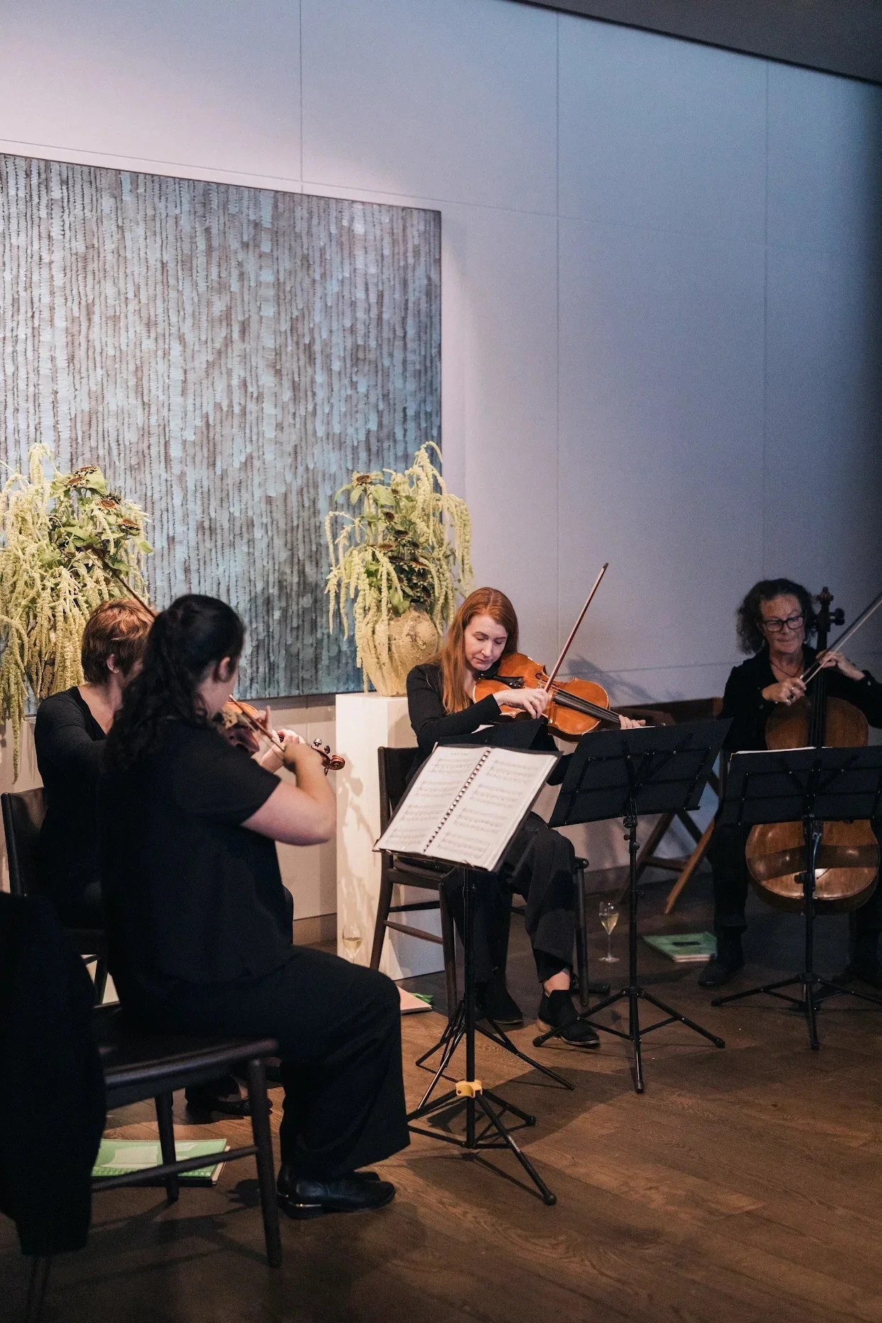 A group of musicians playing string instruments, including violins and a cello, in a performance setting. They are seated with music stands and sheet music in front of them. The background features a large painting or textured artwork and decorative 