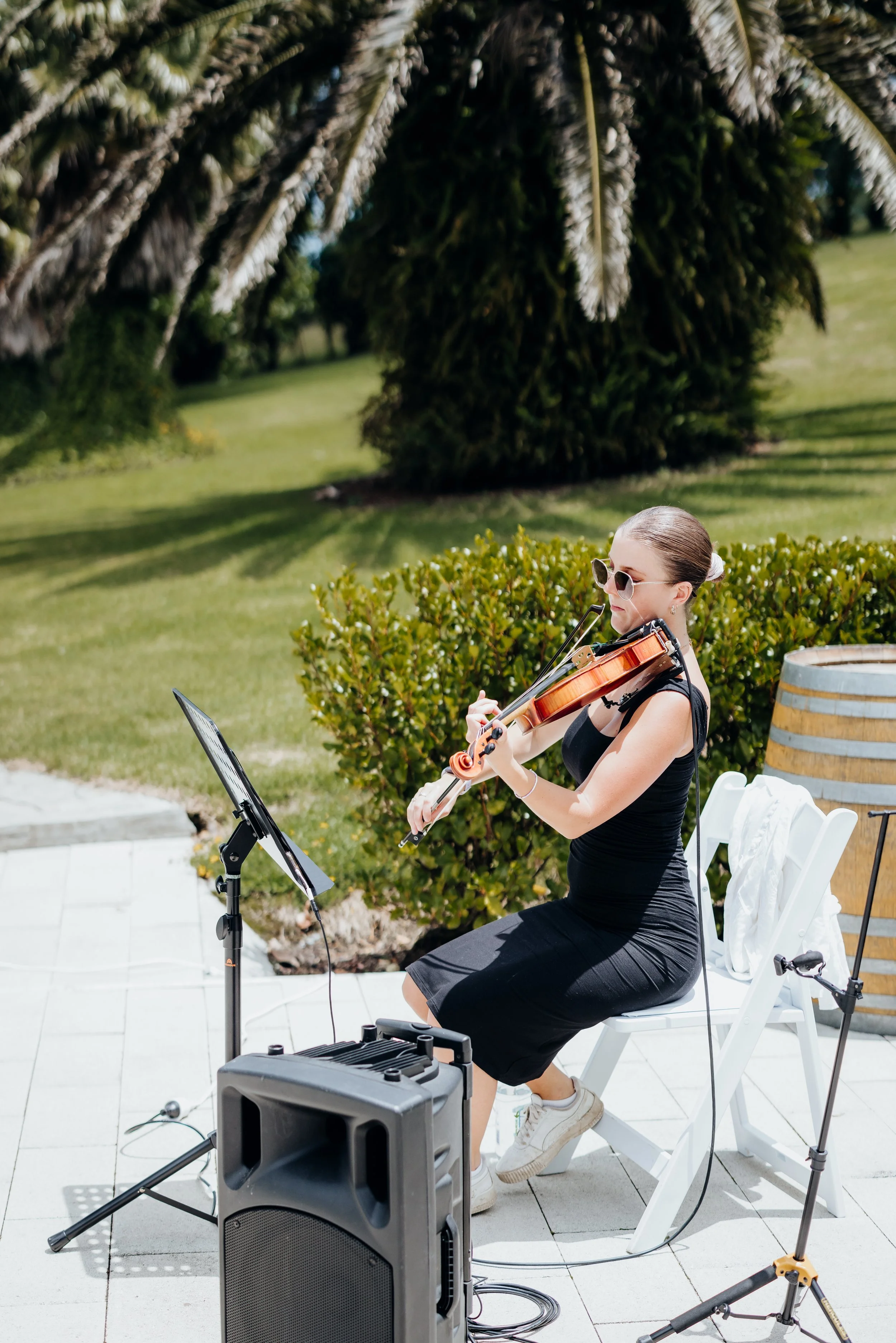 Woman playing a violin outdoors, seated on a white chair, wearing sunglasses and a black dress, with music stand and sound equipment nearby, surrounded by greenery.