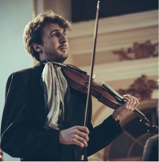Young man playing the violin on stage, dressed in formal attire with a scarf around his neck.