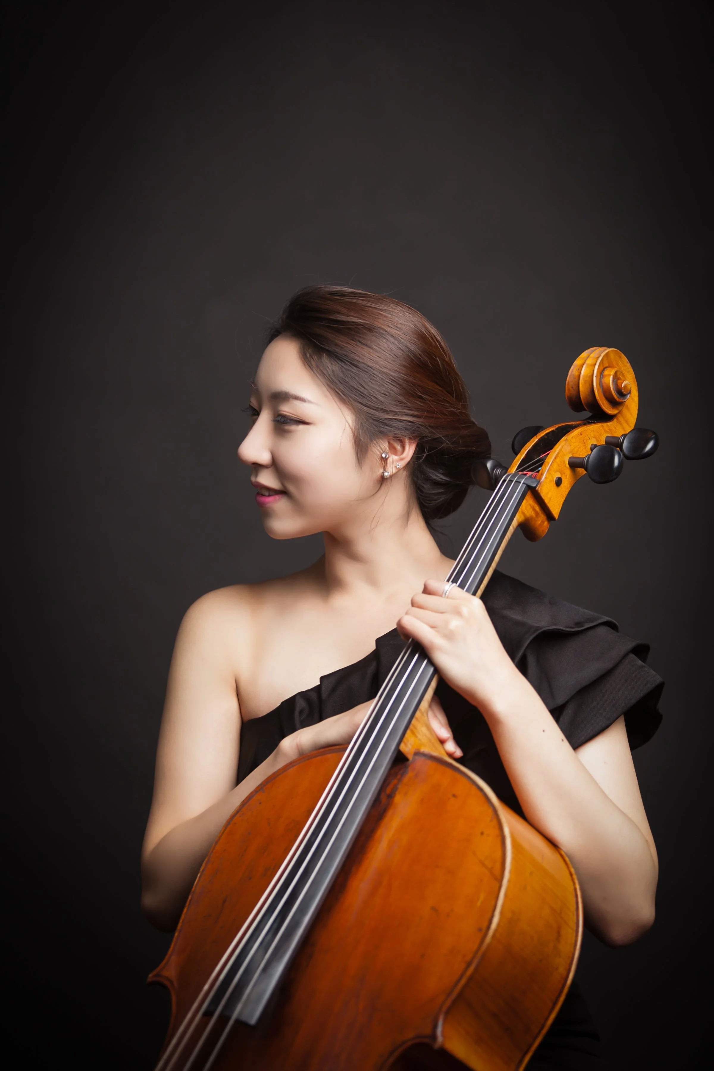 A woman with brown hair styled in a loose updo, wearing a black off-shoulder dress, is holding a wooden cello and appears to be preparing to play or pose with it. The background is dark, highlighting her and the instrument.