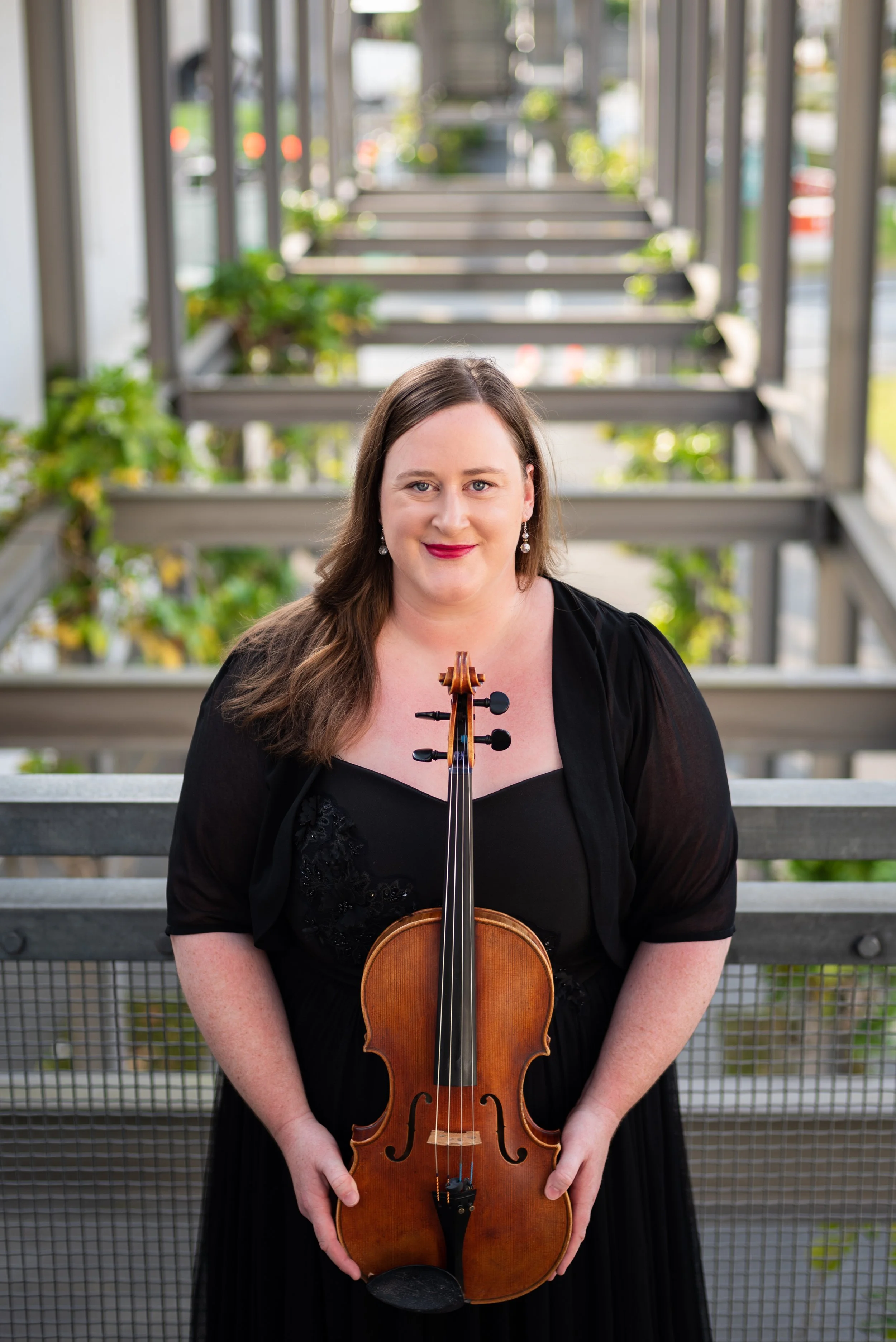 Woman with long brown hair in a black dress holding a violin in front of a metal railing with potted plants on an outdoor balcony.