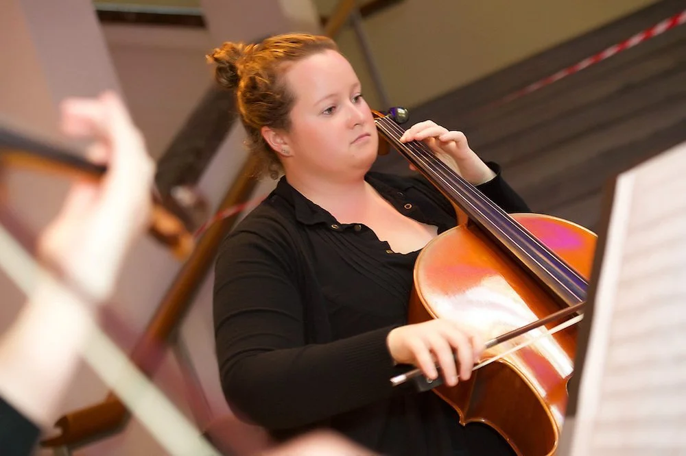 A woman with reddish hair tied back, playing a cello in a music room.