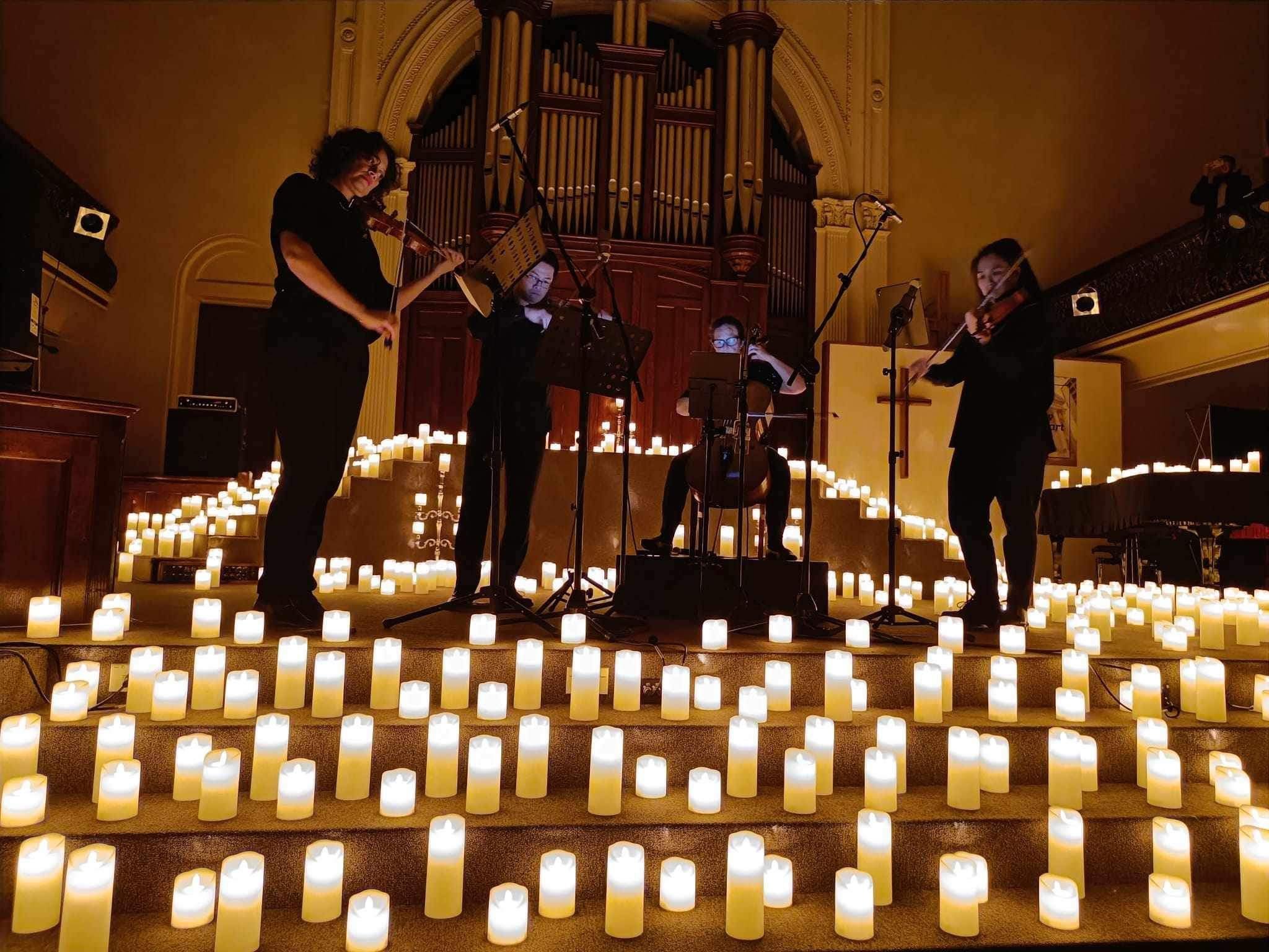 Musicians performing on stage surrounded by numerous candlelights on the floor, with a large organ in the background.