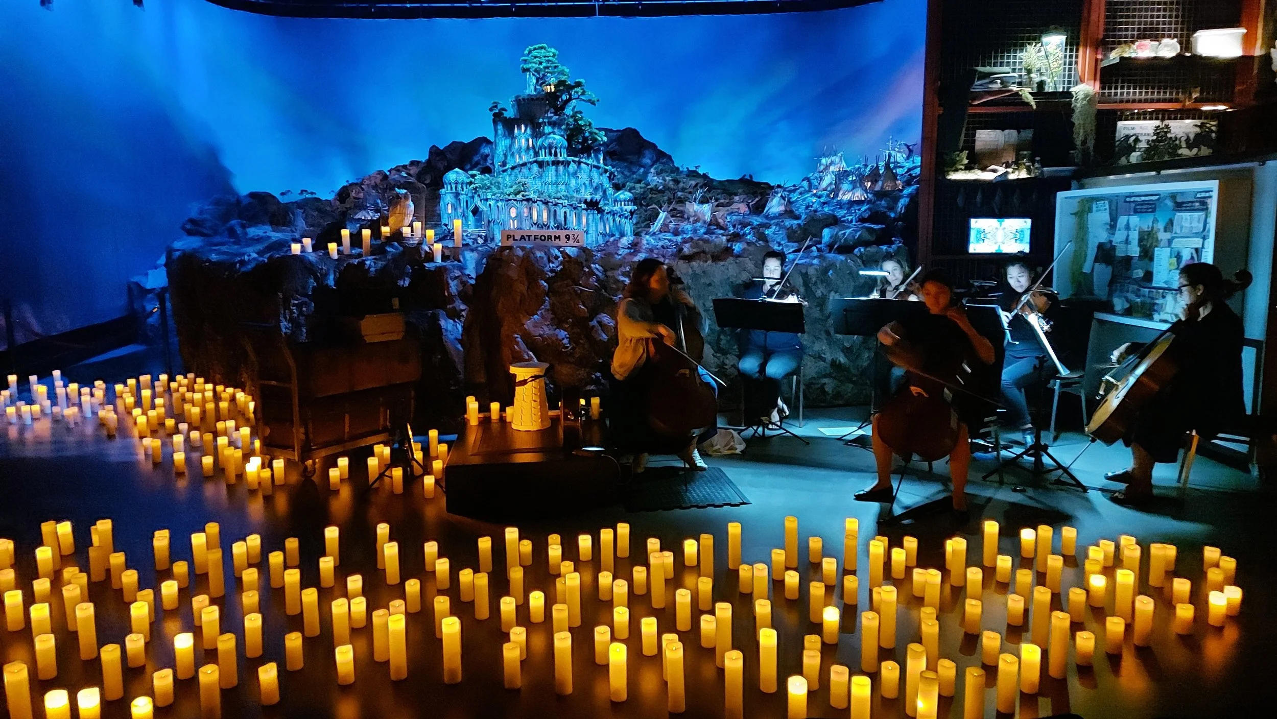 A group of musicians playing string instruments in a dimly lit room decorated with numerous yellow candles. The background features a model of a fantasy castle with a sign that reads 'Platform 9 3/4'.