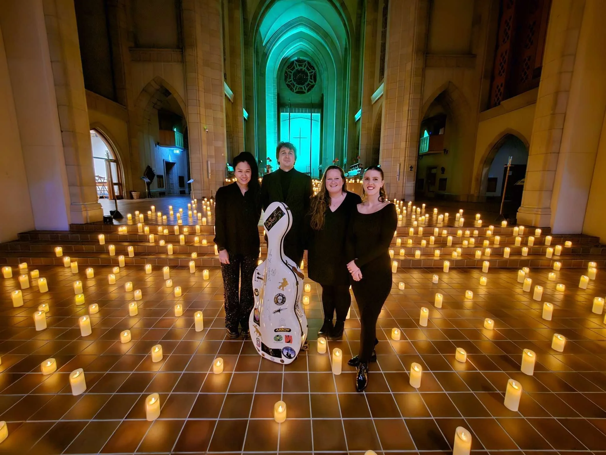 Four people in black attire standing inside a church or cathedral decorated with numerous lit candles on the floor. The background features a large stained glass window and arches illuminated with colorful lights.