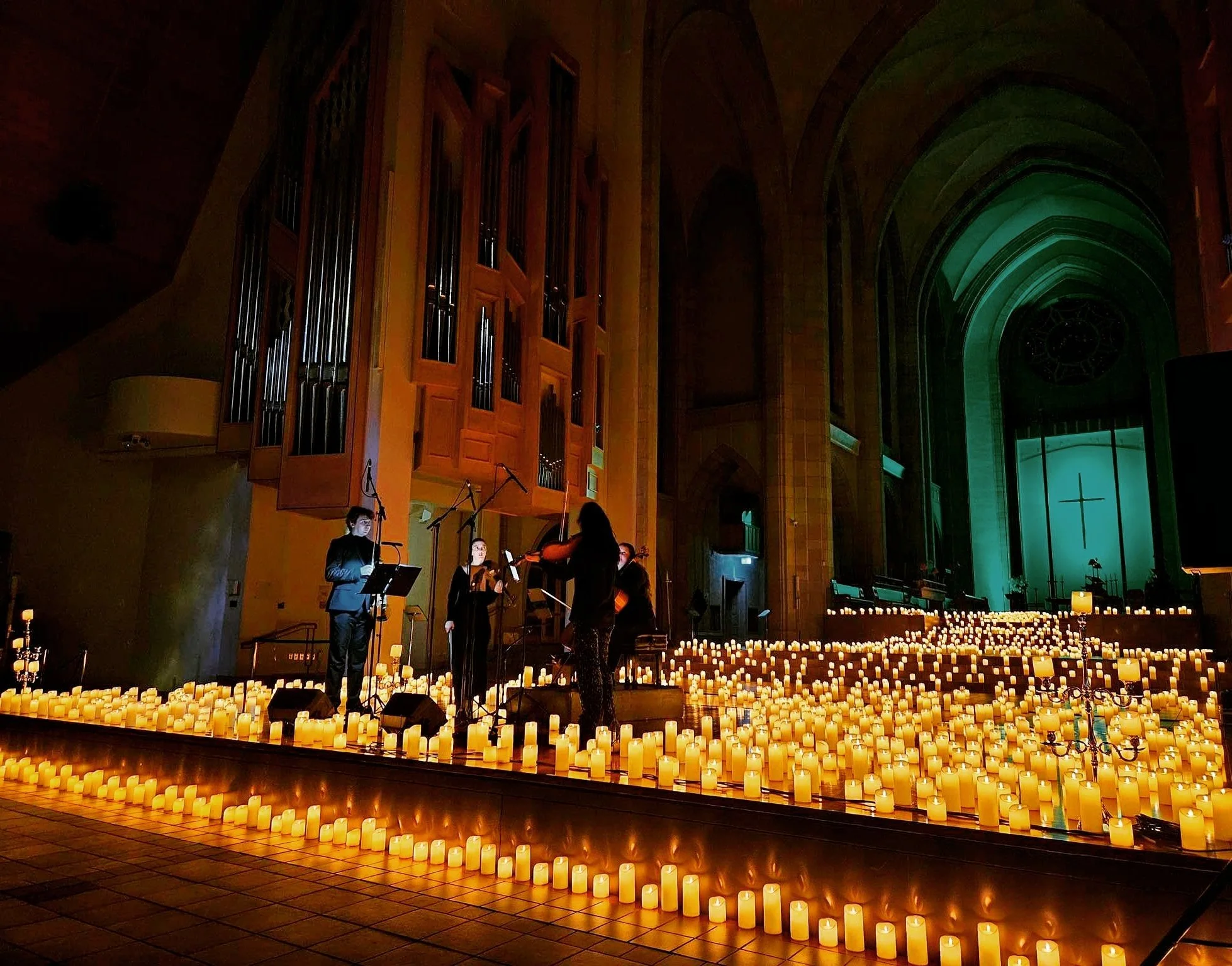 A group of people performing a musical concert in a church, illuminated by numerous candles on the floor, with an organ and large arch window in the background.