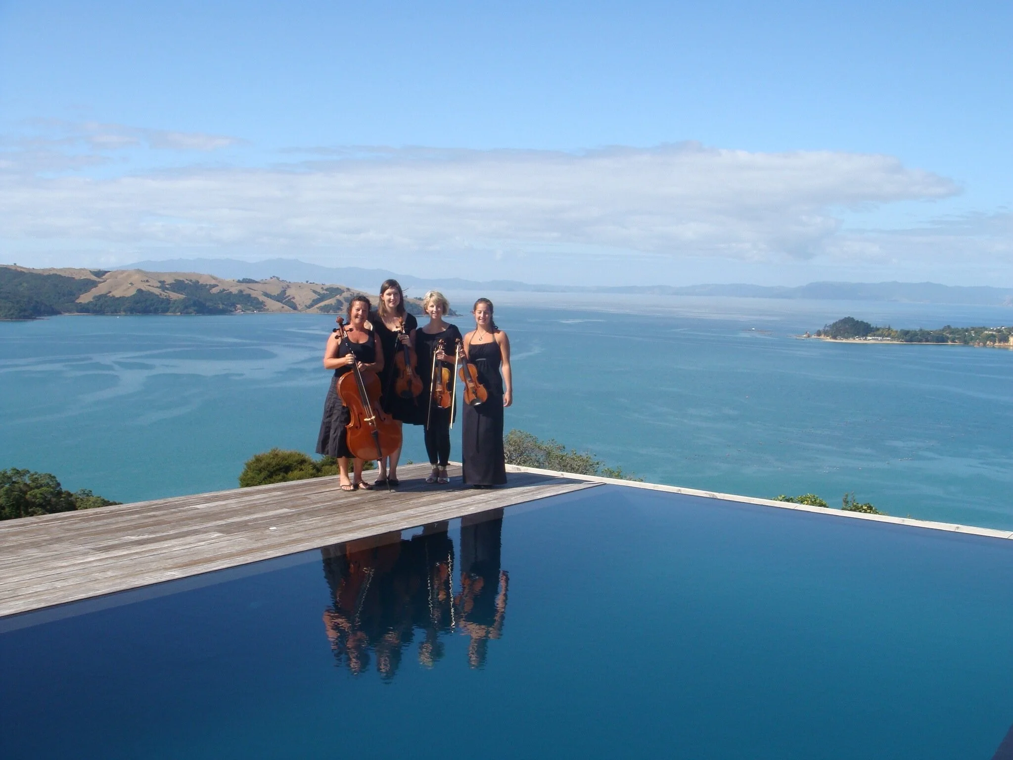 Four women standing on a wooden terrace holding musical instruments, with a scenic body of water and rolling hills in the background.