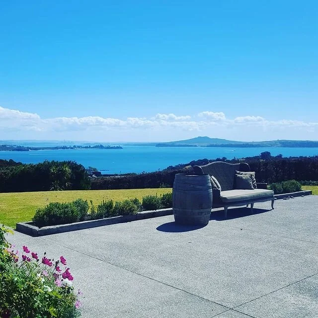 A scenic outdoor patio with a sofa and a barrel-shaped side table, overlooking a body of water with mountains and a partly cloudy sky in the background.