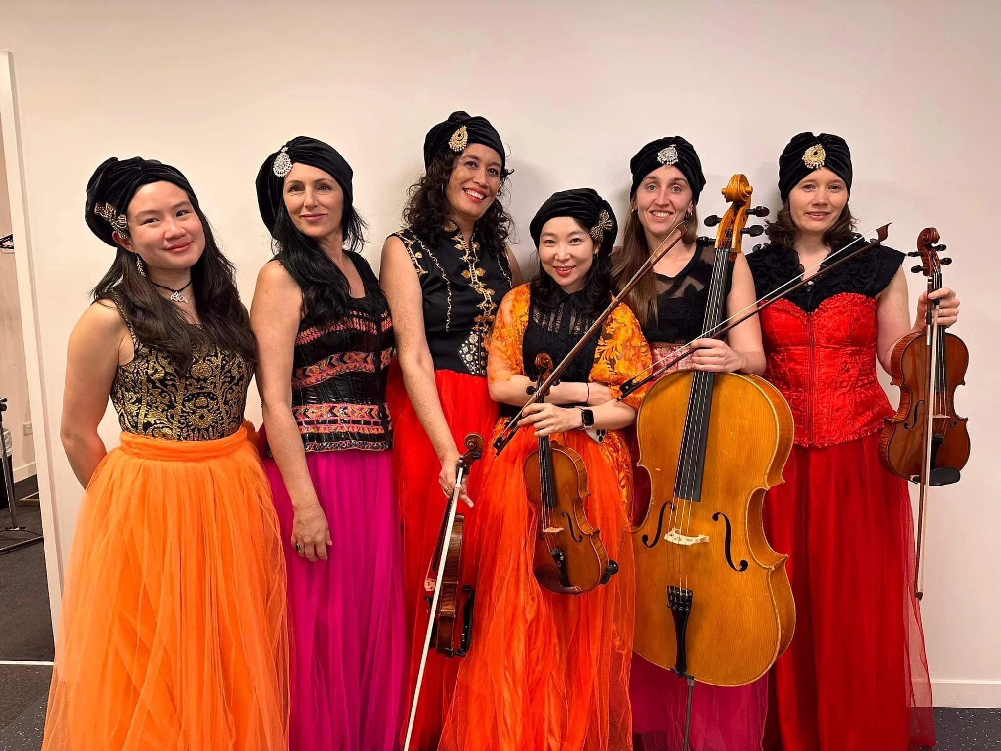 Group of seven women dressed in colorful ethnic outfits and black headwraps, holding musical instruments including violins and cellos, standing against a plain light-colored wall.