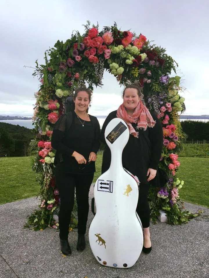 Two women stand in front of a large floral arch, smiling at the camera. One woman has dark hair and is wearing a black outfit with a backpack, the other has curly hair, is wearing a black top with a pink scarf, and holds a small bag. A white golf clu