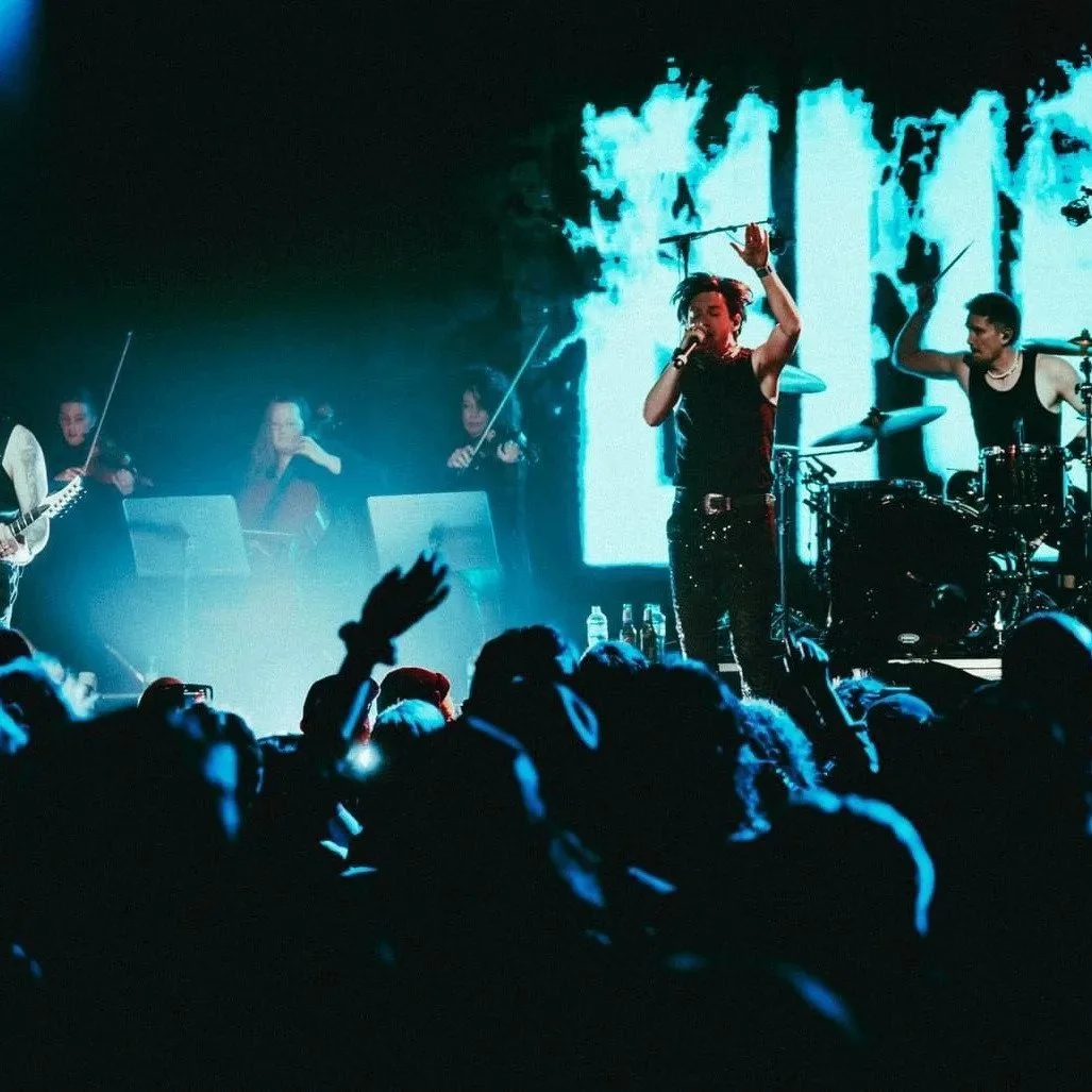 Performers on stage during a concert, with a singer holding a microphone and a drummer playing, and a large screen in the background displaying blue graphics, while an audience watches and raises hands.