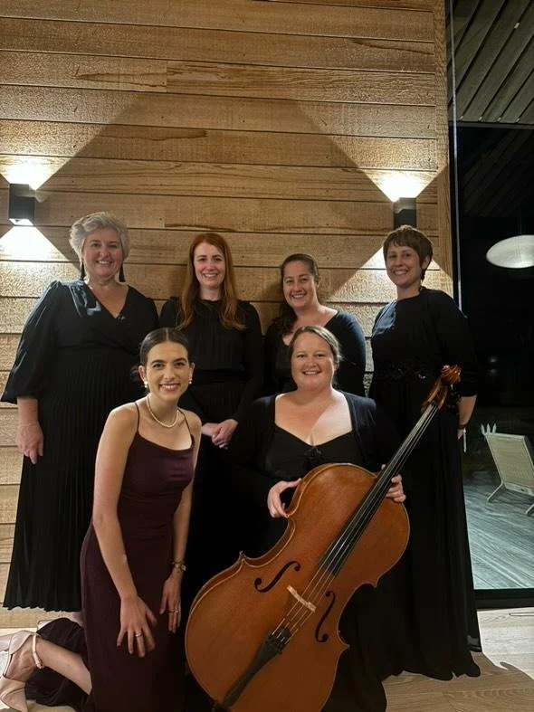 Six women dressed in formal black attire, with one woman holding a cello, standing in front of a wooden wall with modern lighting.