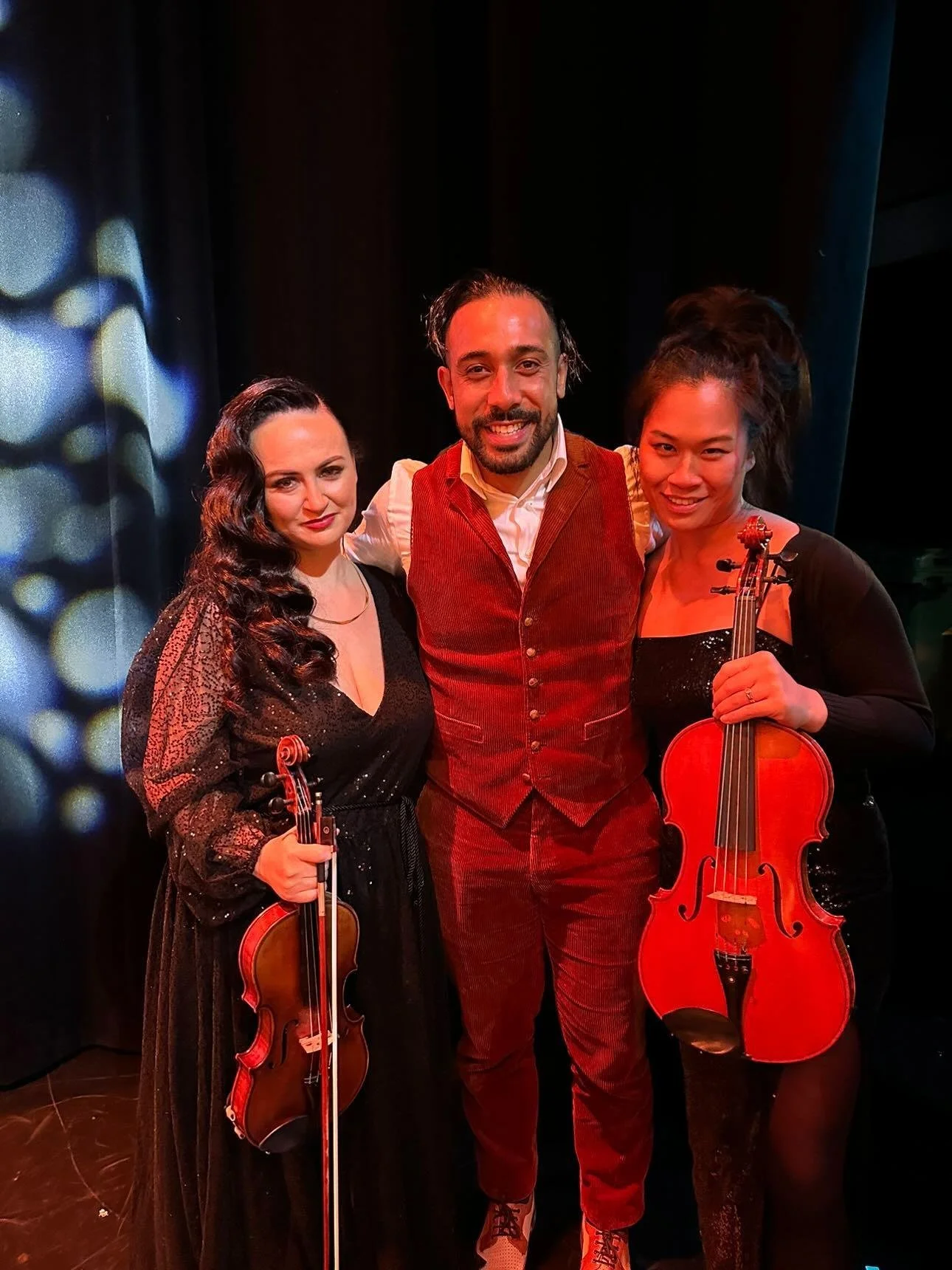 Three musicians, two women and one man, standing together after a performance, holding violins, on a stage with a dark background.