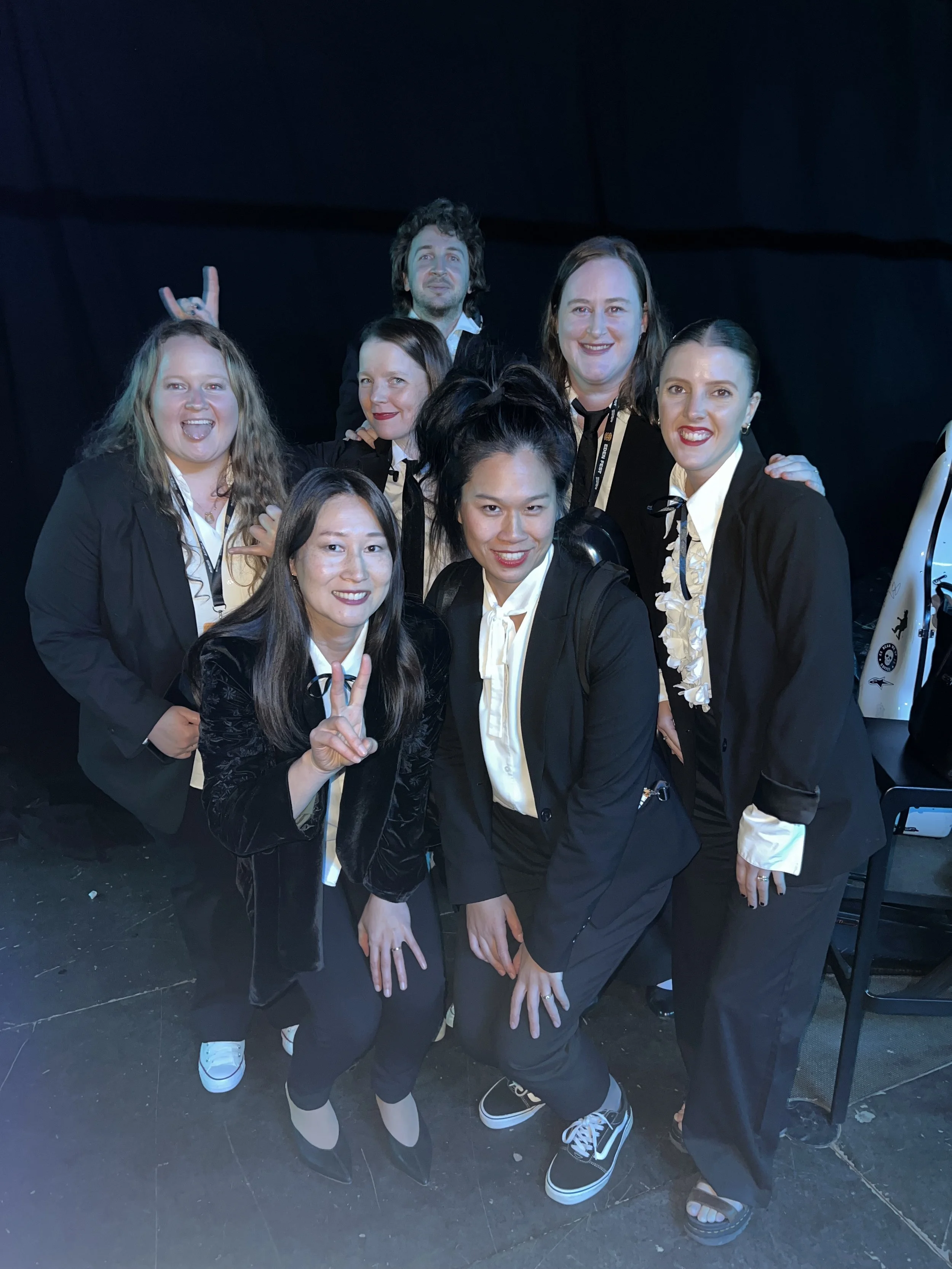 Group of seven people dressed in black suits with white shirts, posing together against a dark background. Some are making peace signs or playful gestures, smiling at the camera.
