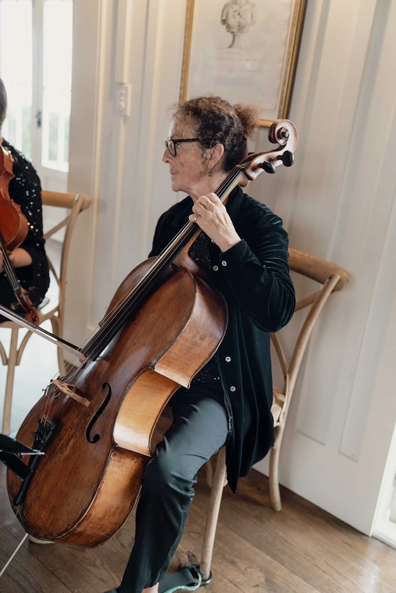 An elderly woman wearing glasses playing a cello indoors.
